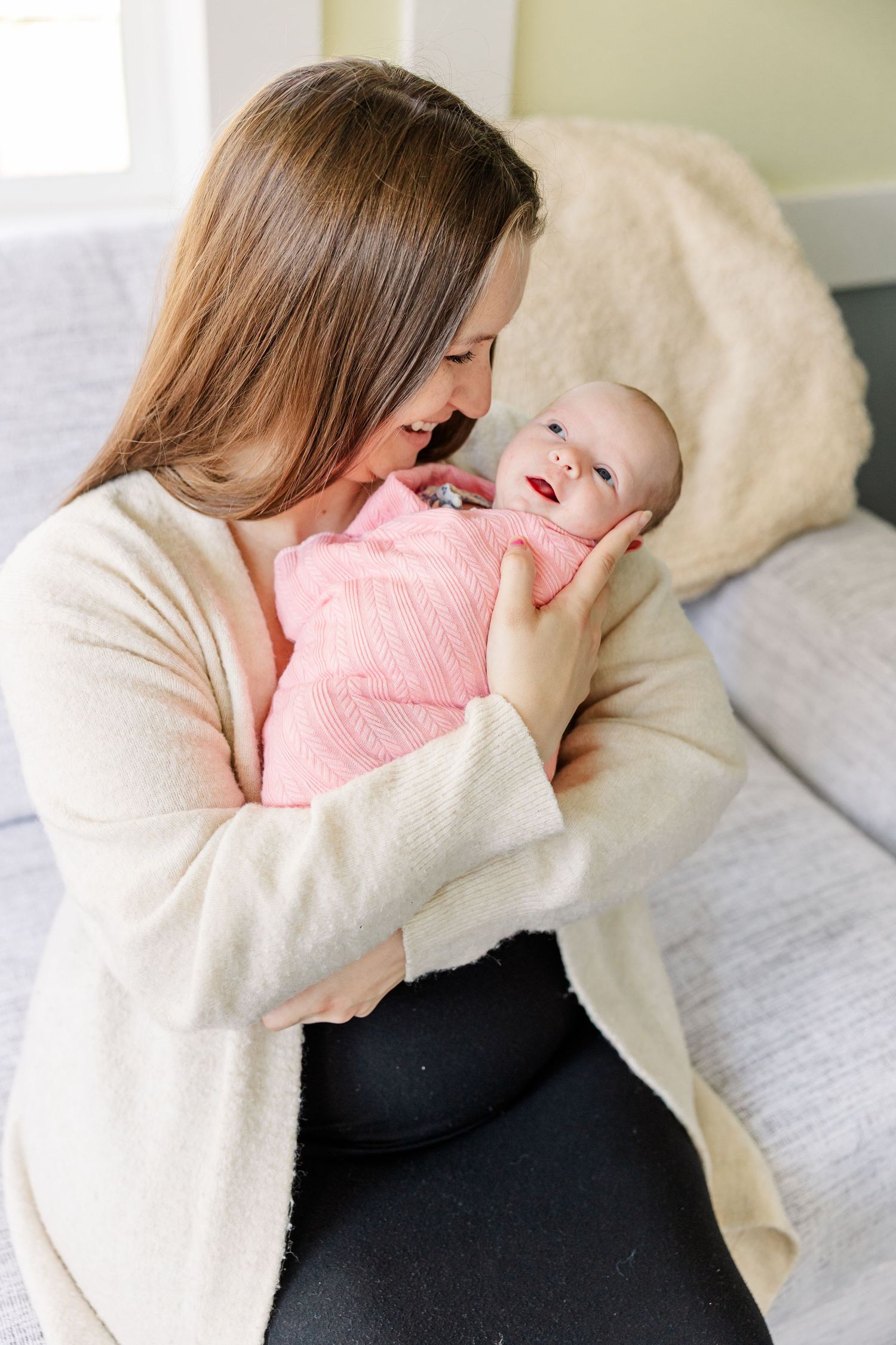 A loving mother in a cream cardigan gazes at her newborn baby wrapped in a pink swaddle while sitting on a couch.