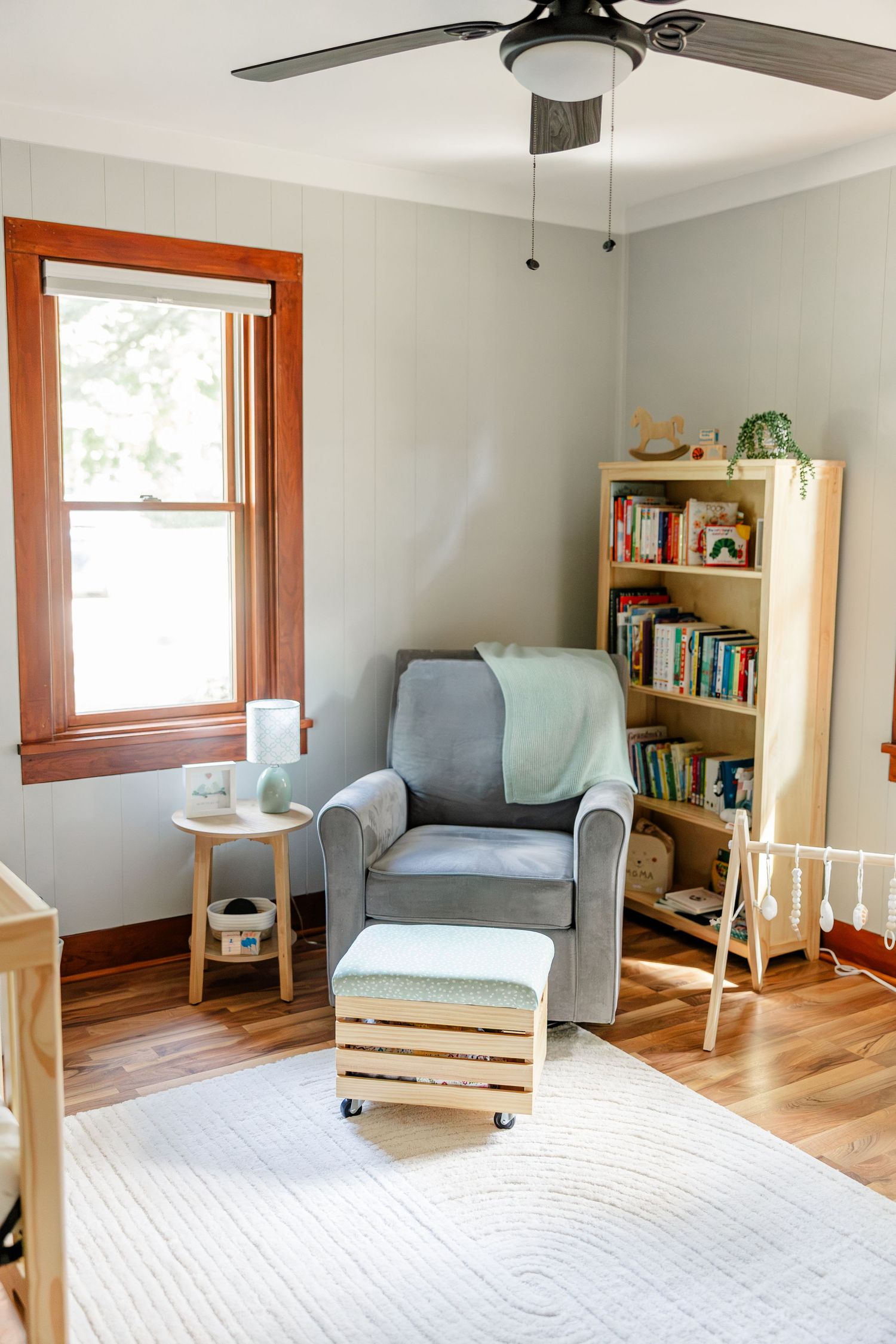 Cozy nursery corner featuring a gray glider chair, wooden bookshelf, and natural wood accents with soft lighting.
