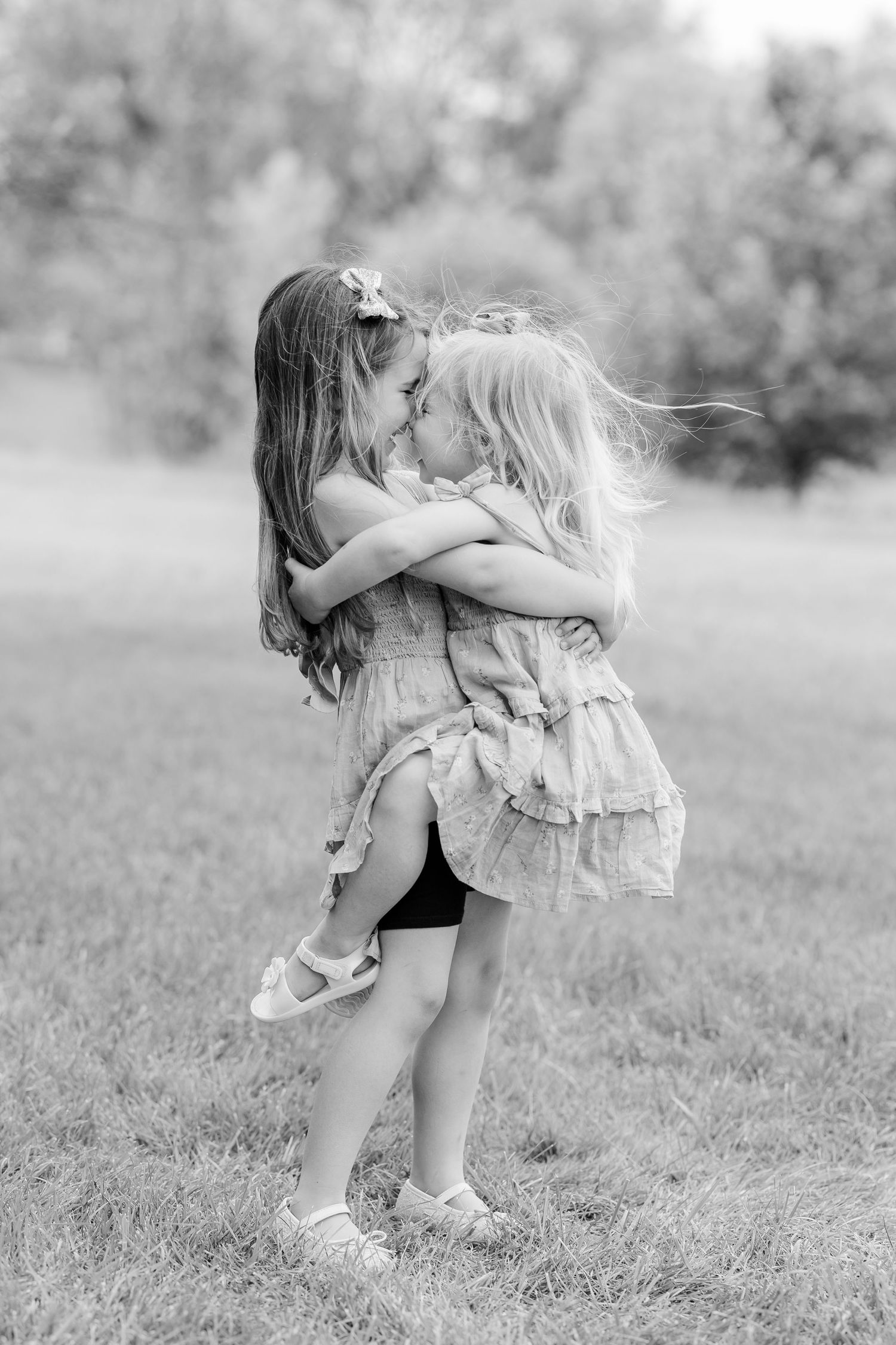 Two young girls in ruffled dresses embrace and touch noses in a sweet moment captured in black and white.
