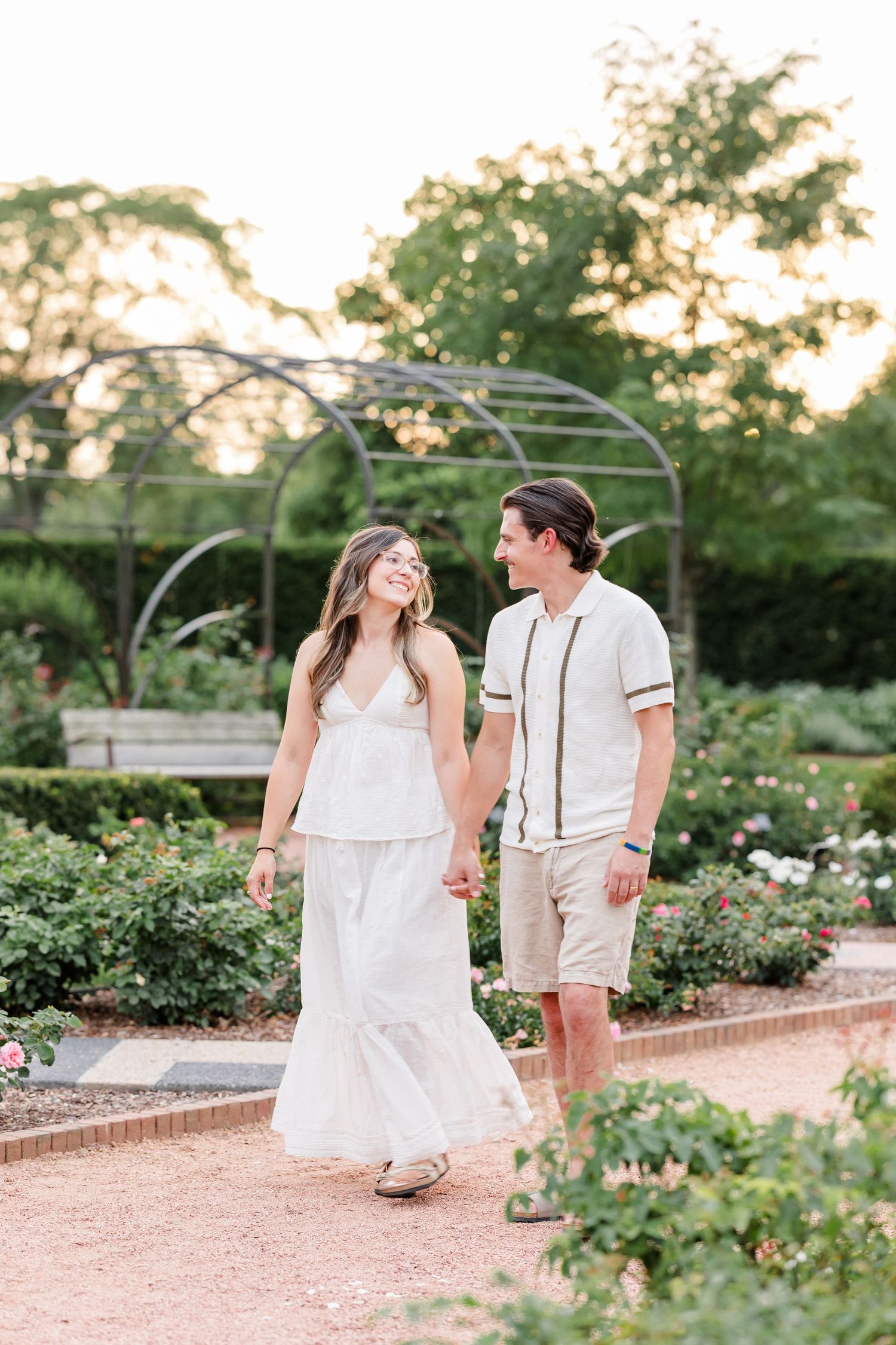 Couple in white outfits walk hand in hand through rose garden at golden hour sunset.