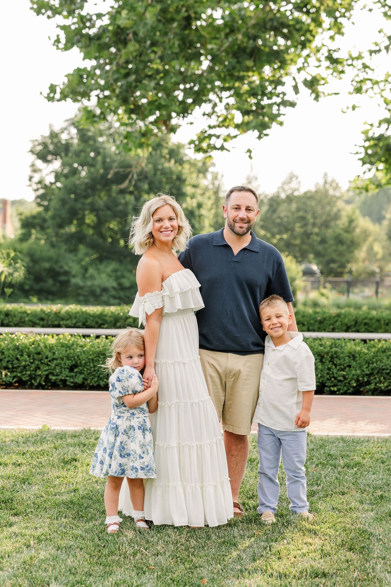 Happy family of four poses on green lawn with lush garden backdrop during golden hour outdoor portrait session.
