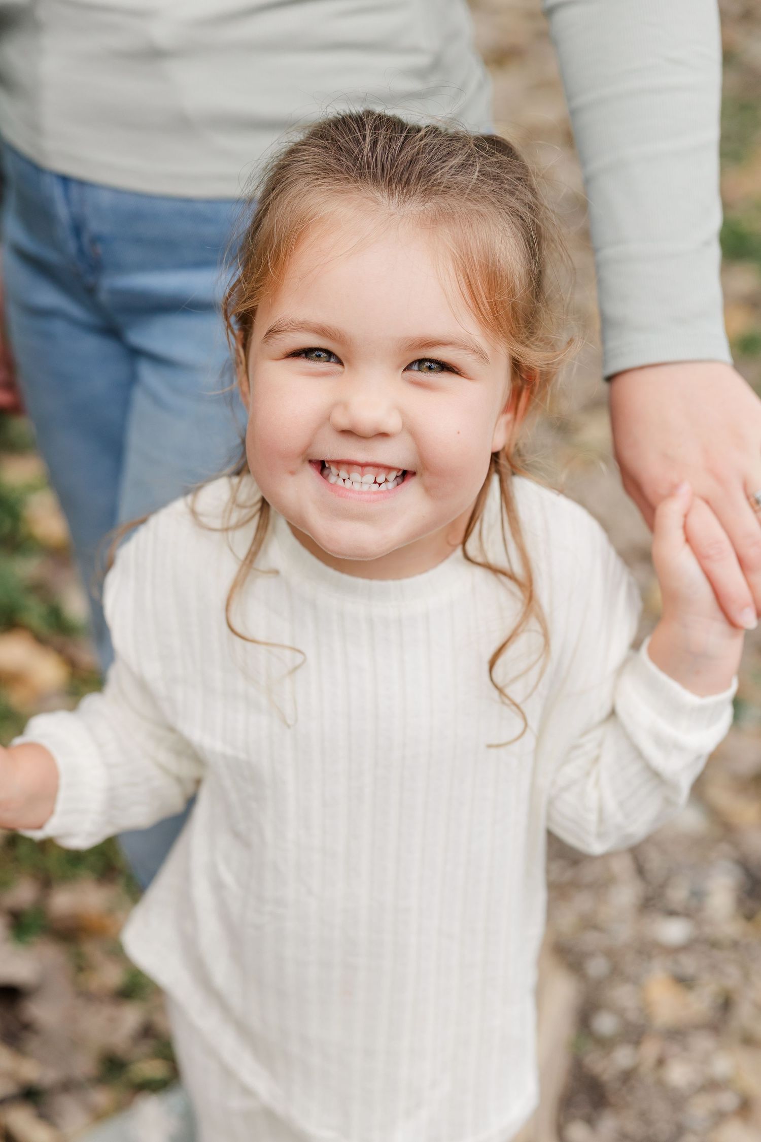 A joyful young girl in a white ribbed sweater smiles brightly while holding an adult's hand outdoors.