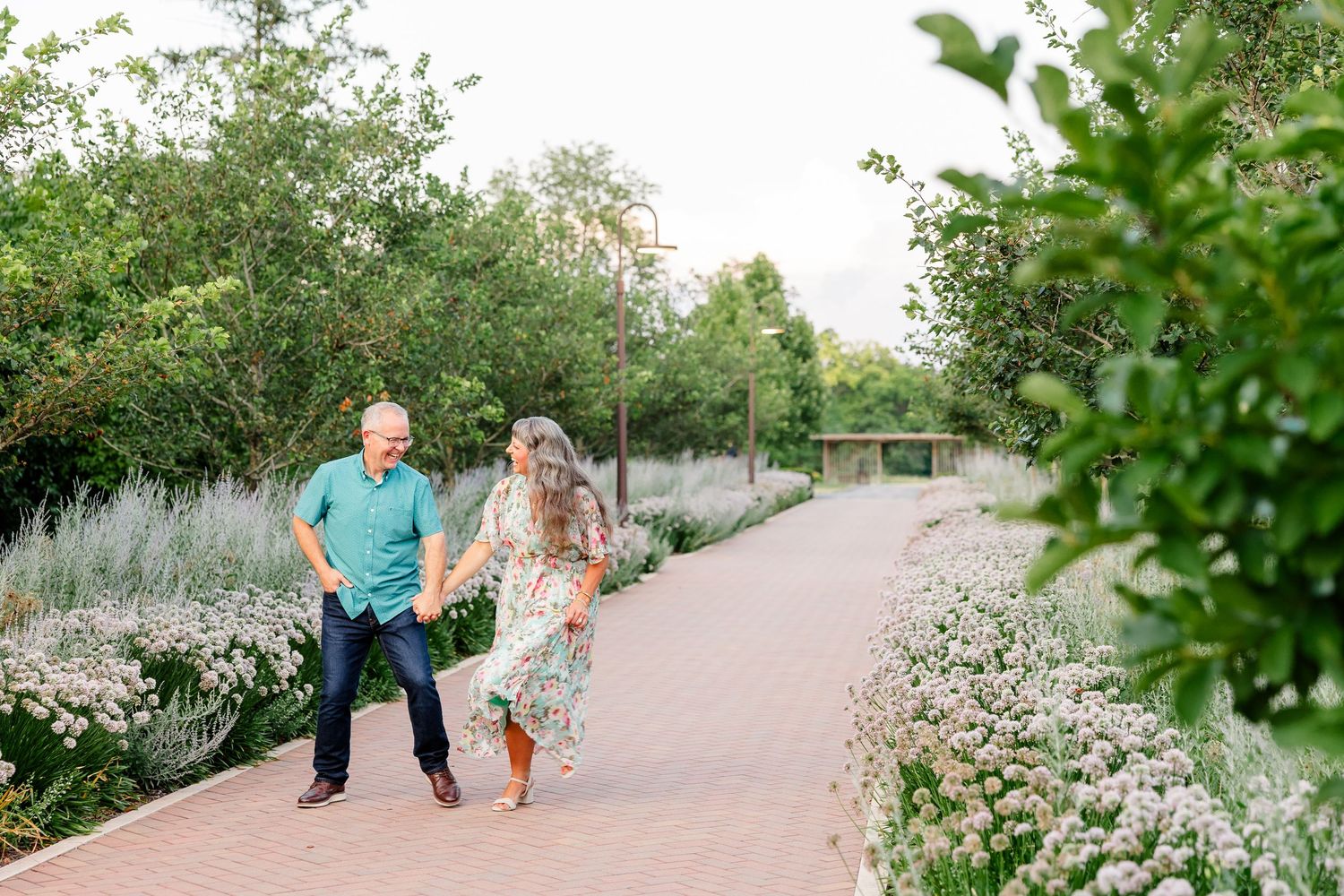 Happy couple holding hands and walking along a brick garden path surrounded by white flowers.