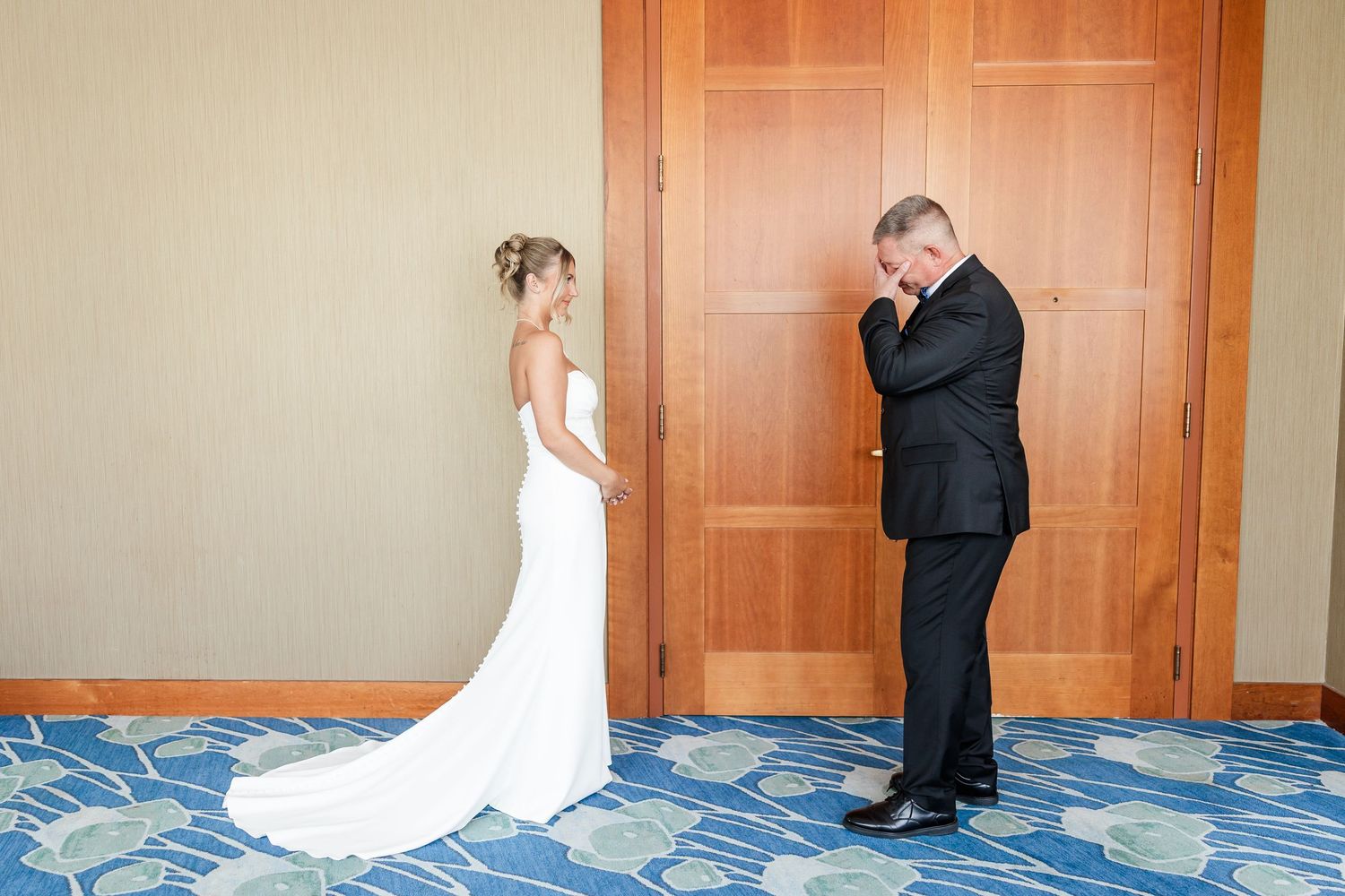 Emotional groom wiping tears during first look with bride in white wedding gown before wooden doors.
