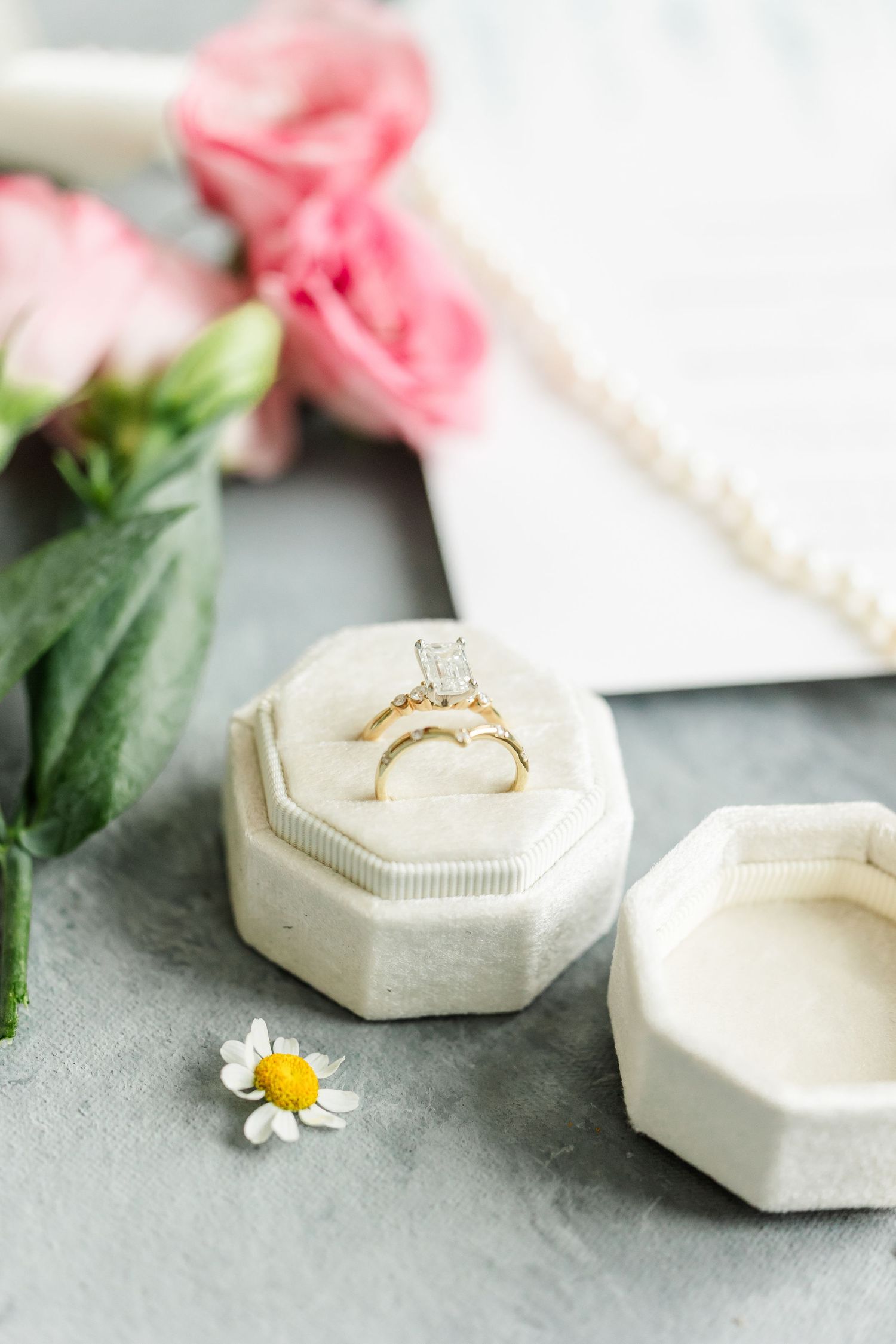 Gold diamond engagement ring in white velvet octagon box with pink roses and daisy on gray surface.