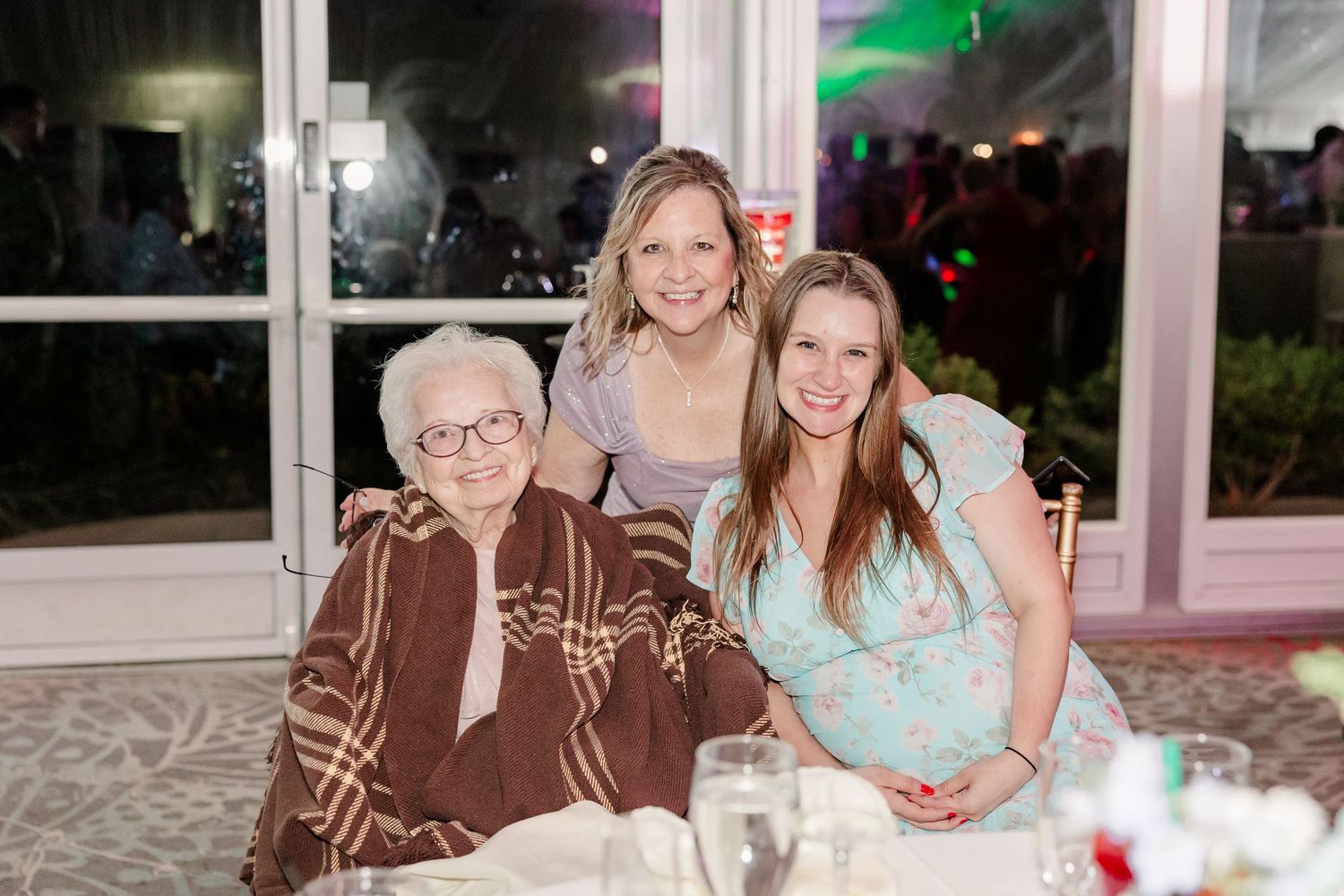 Three generations of women smiling together at wedding reception dinner table at night.