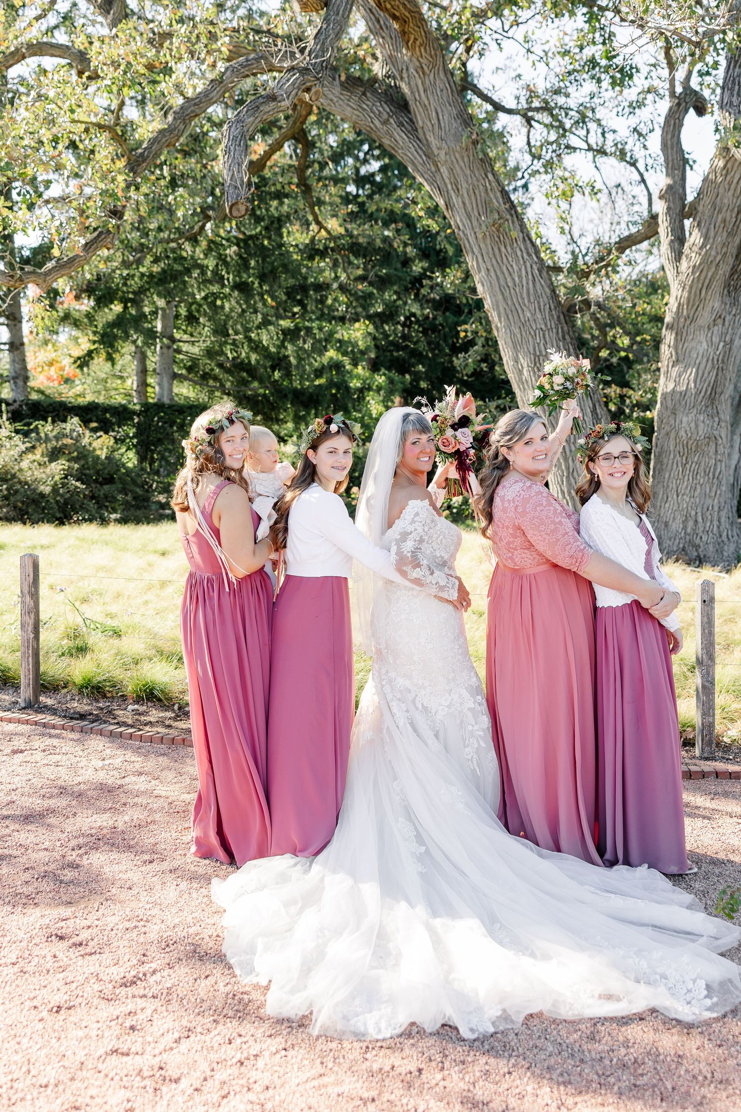 Bride in a lace wedding gown with a long train poses with four bridesmaids in dusty rose dresses under a large tree outdoors.