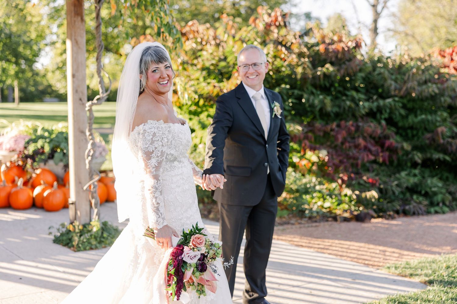 Smiling bride in a lace gown and veil holding a floral bouquet walks hand in hand with her groom in a dark suit near autumn pumpkins.