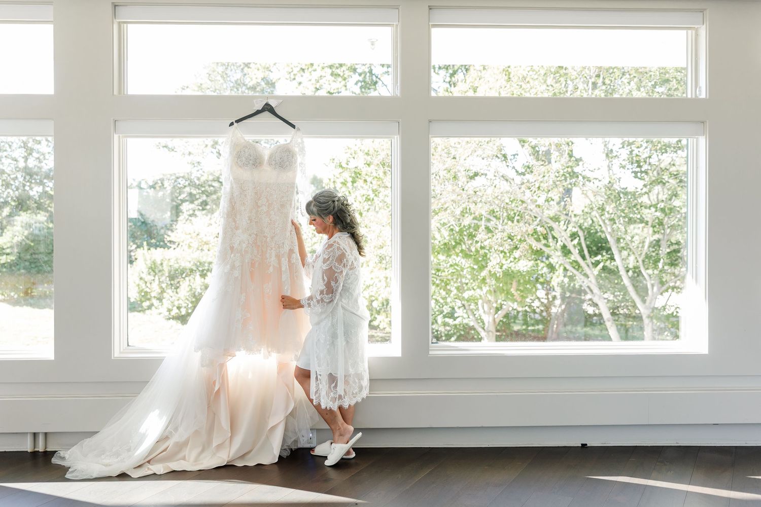 A bride in a lace robe stands beside her elegant wedding dress hanging in front of bright windows overlooking green trees.