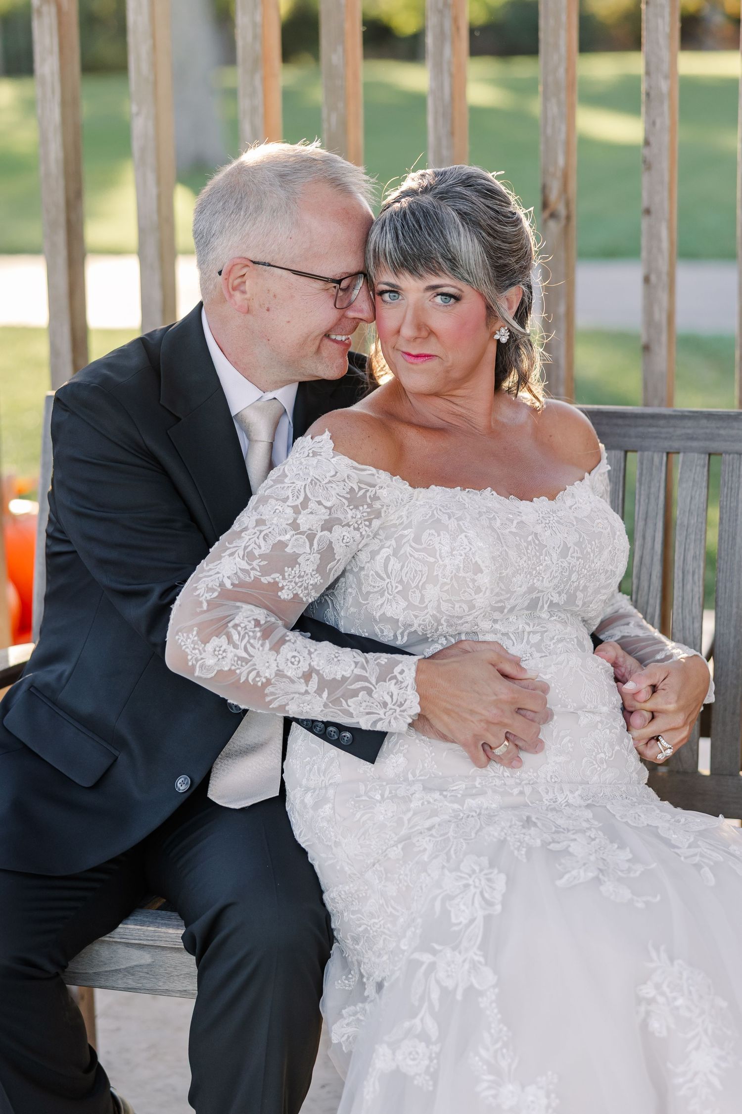 Groom in a dark suit and glasses rests his forehead against his bride in a lace wedding dress as they sit on a wooden bench.