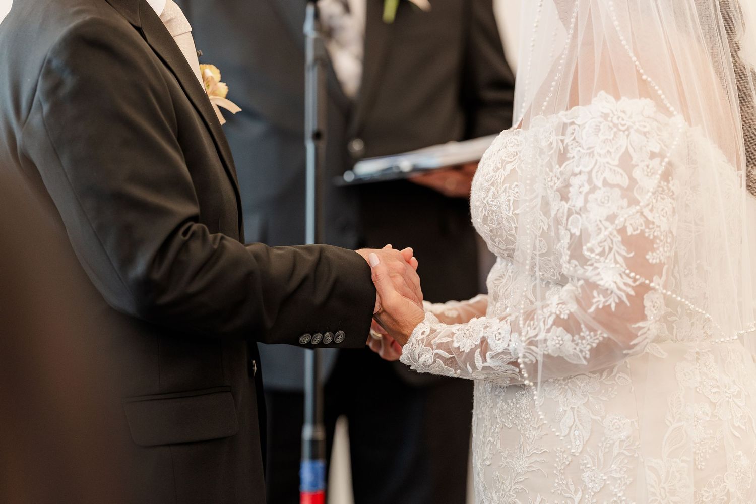 A bride in a lace wedding dress and veil holds hands with a groom in a dark suit as an officiant stands behind them.