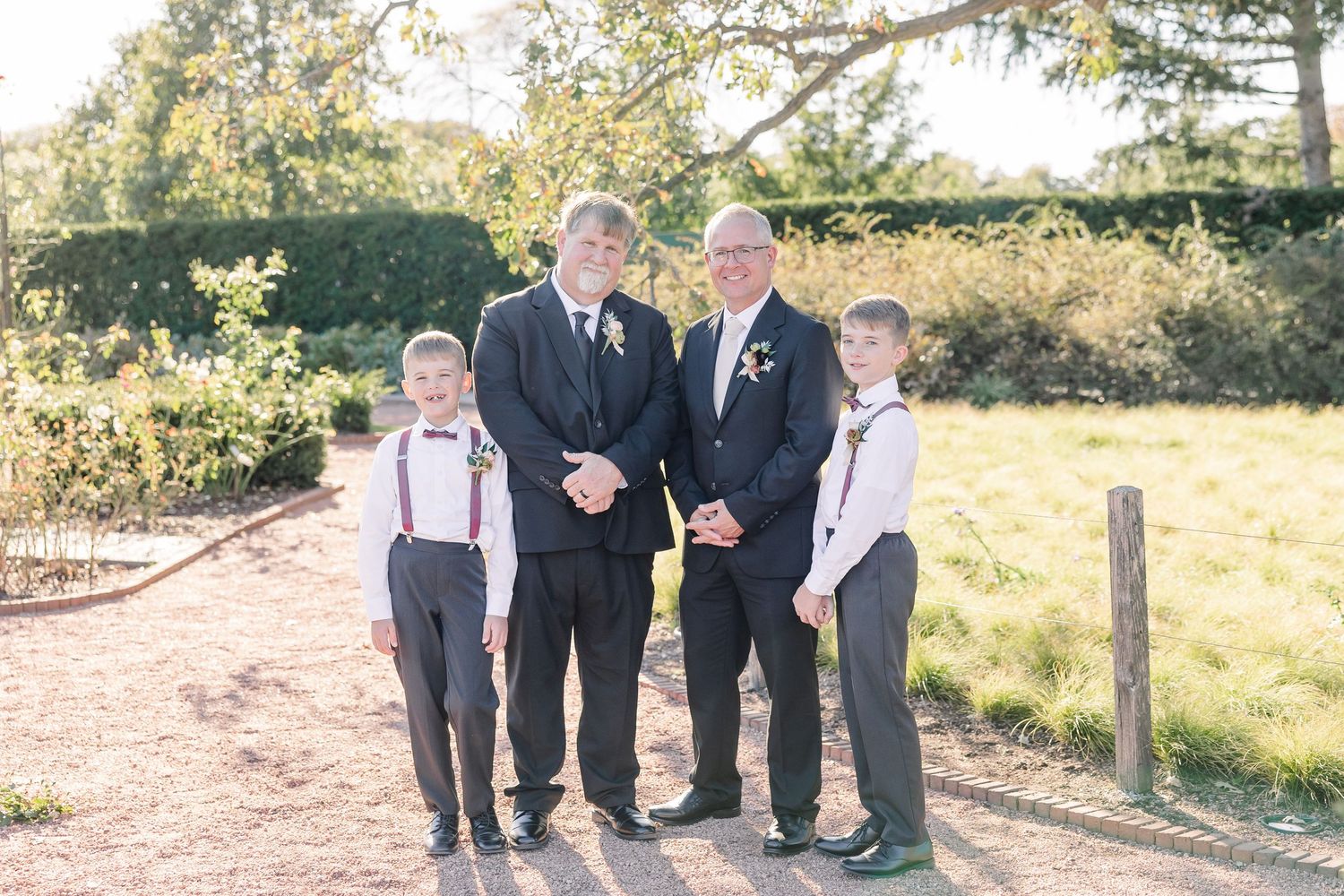 Two men in dark suits with boutonnieres stand beside two boys in suspenders and bow ties on a garden path at an outdoor wedding.