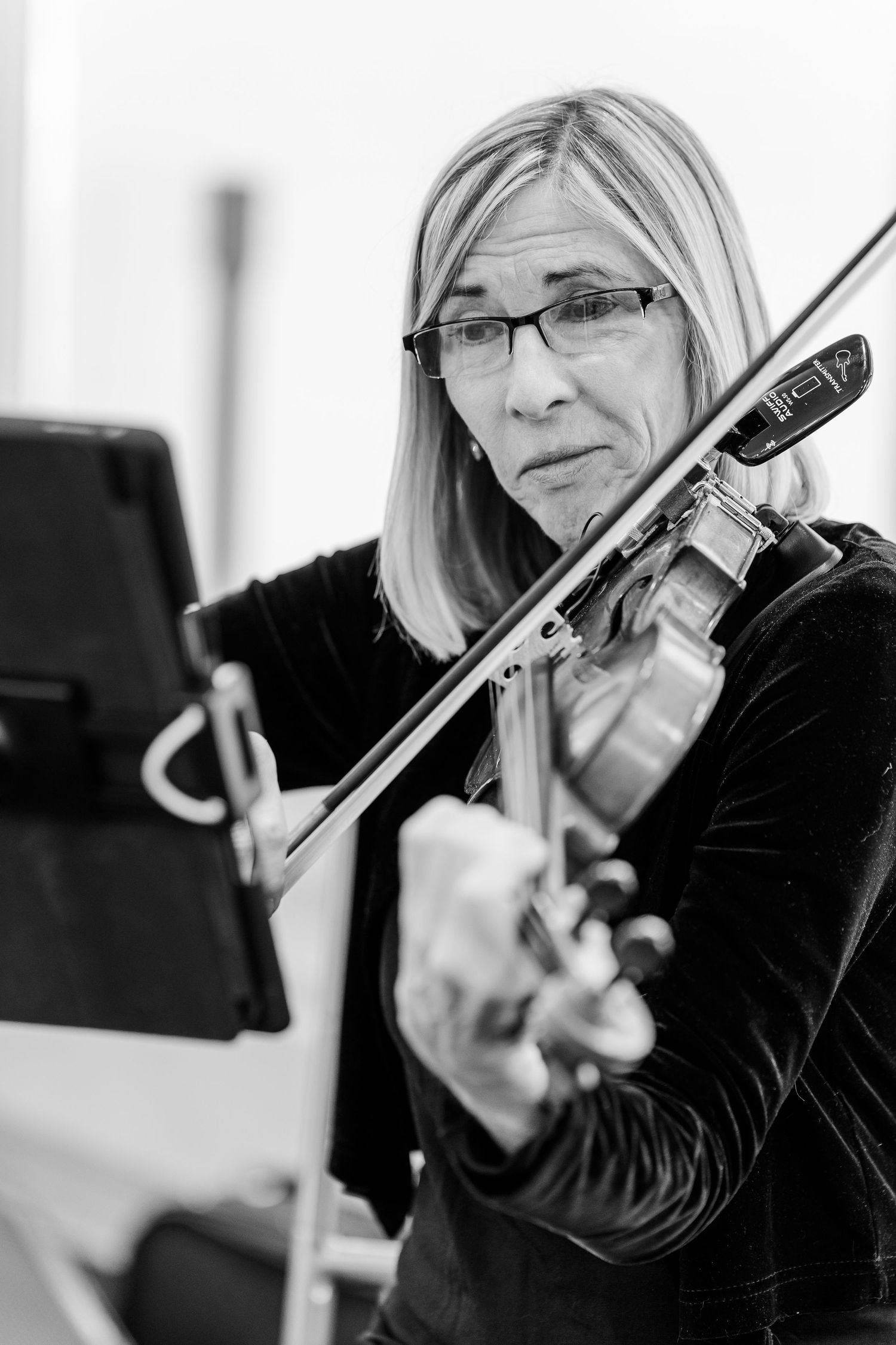 Black and white portrait of a woman with glasses and shoulder-length hair playing the violin beside a music stand.