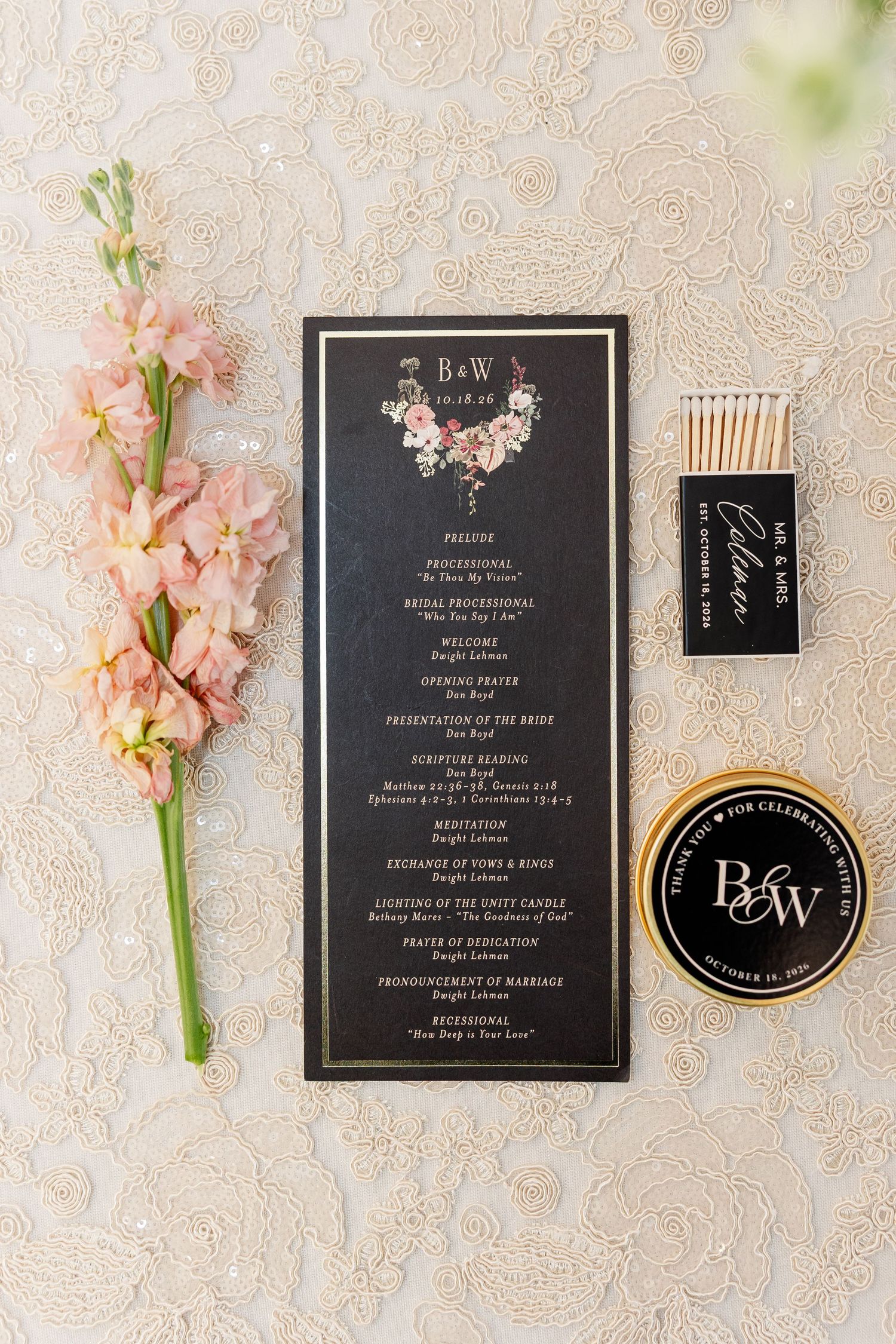 Black and gold wedding ceremony program with floral crest beside pink stock flowers, monogrammed matchbook and B&W round favor on lace.