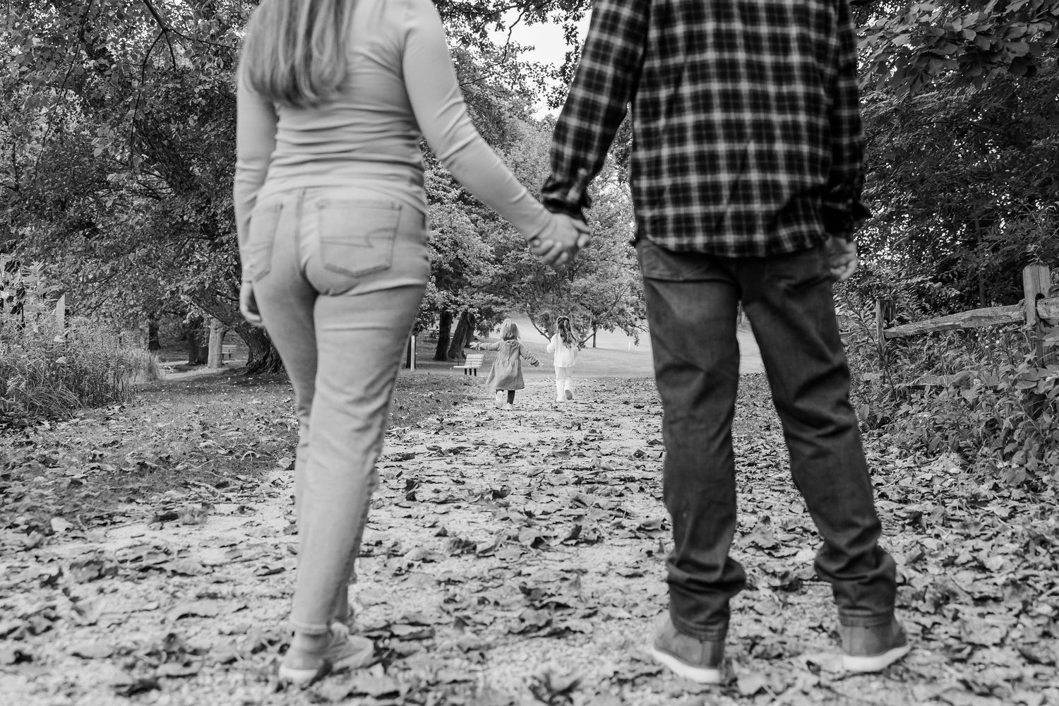 Black and white photo of a couple holding hands walking on a leaf-covered path in autumn.