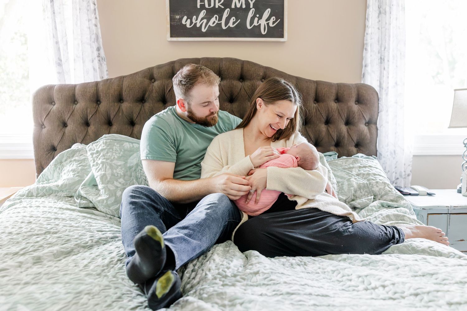A couple sits on a bed, lovingly cradling their newborn baby in a cozy bedroom setting.
