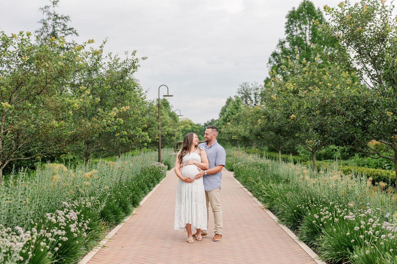 Wide portrait of expecting parents standing on a tree-lined garden path at Cantigny Park