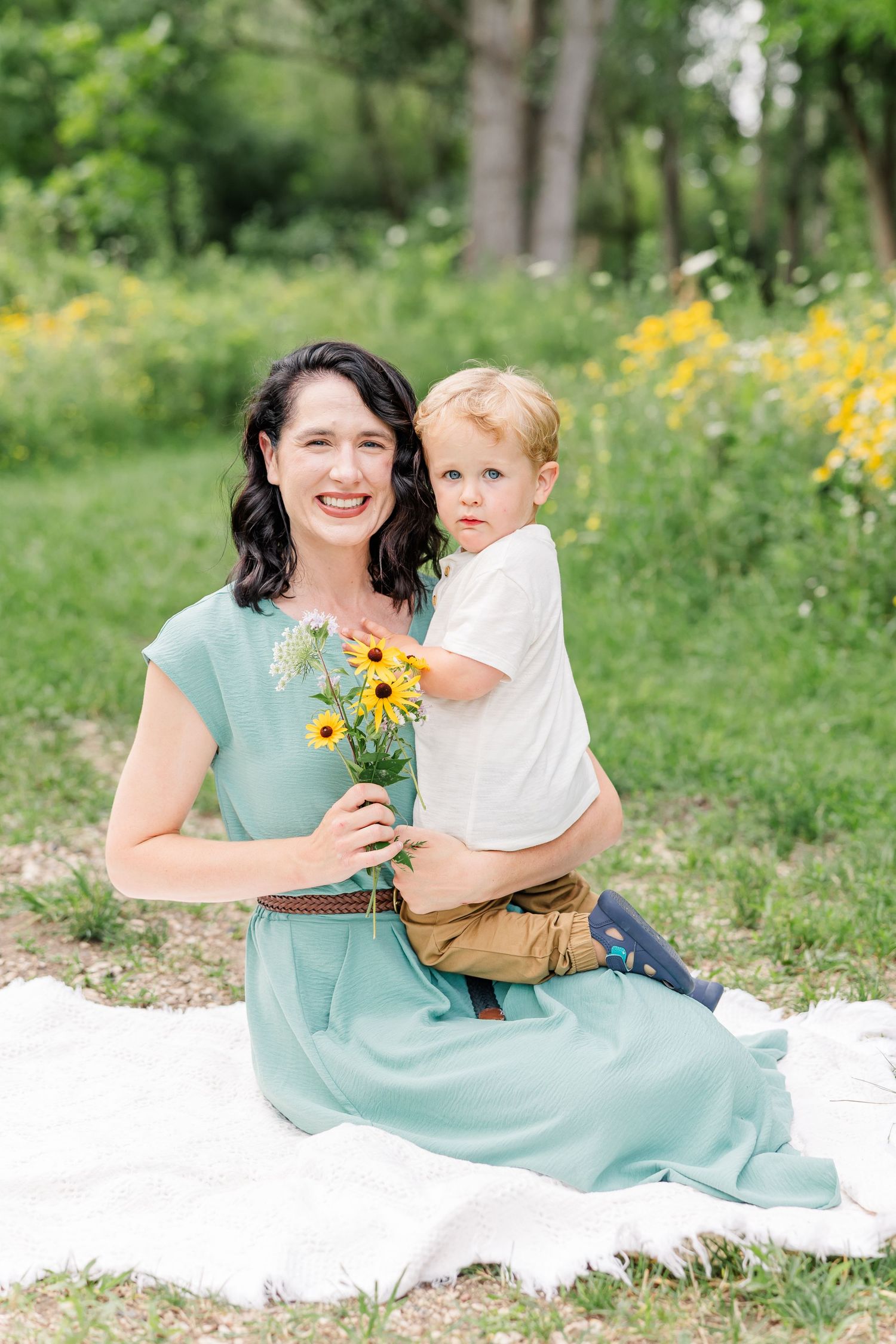 A smiling woman in a teal dress sits on a blanket outdoors holding sunflowers while a toddler boy stands behind her.