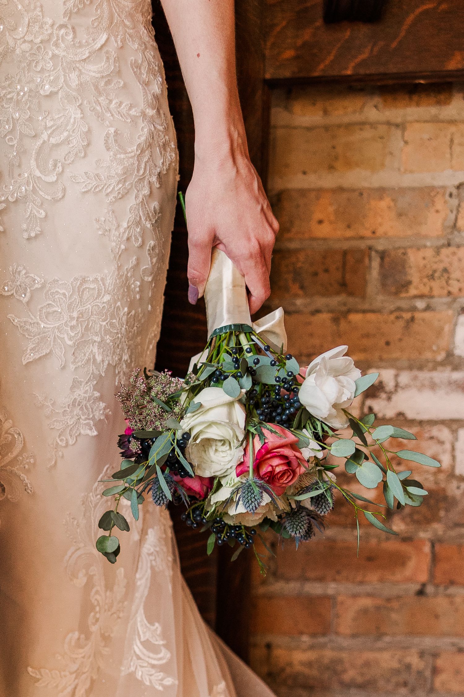 A delicate bridal bouquet with white and pink roses against a lace wedding dress and brick wall.