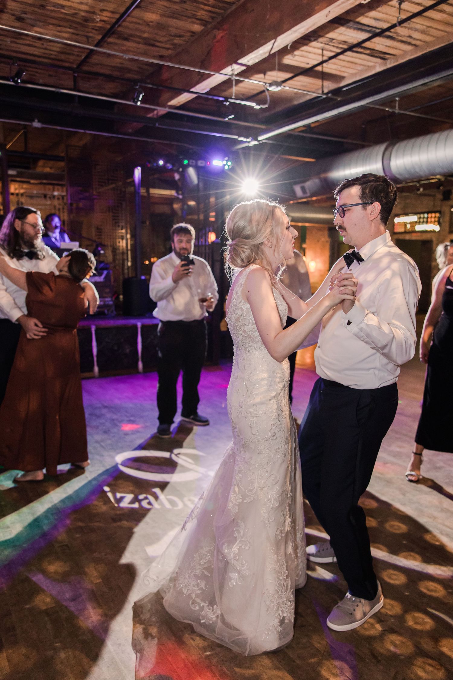 A couple dances in an industrial-style venue with exposed ceiling and purple lighting during a wedding reception.