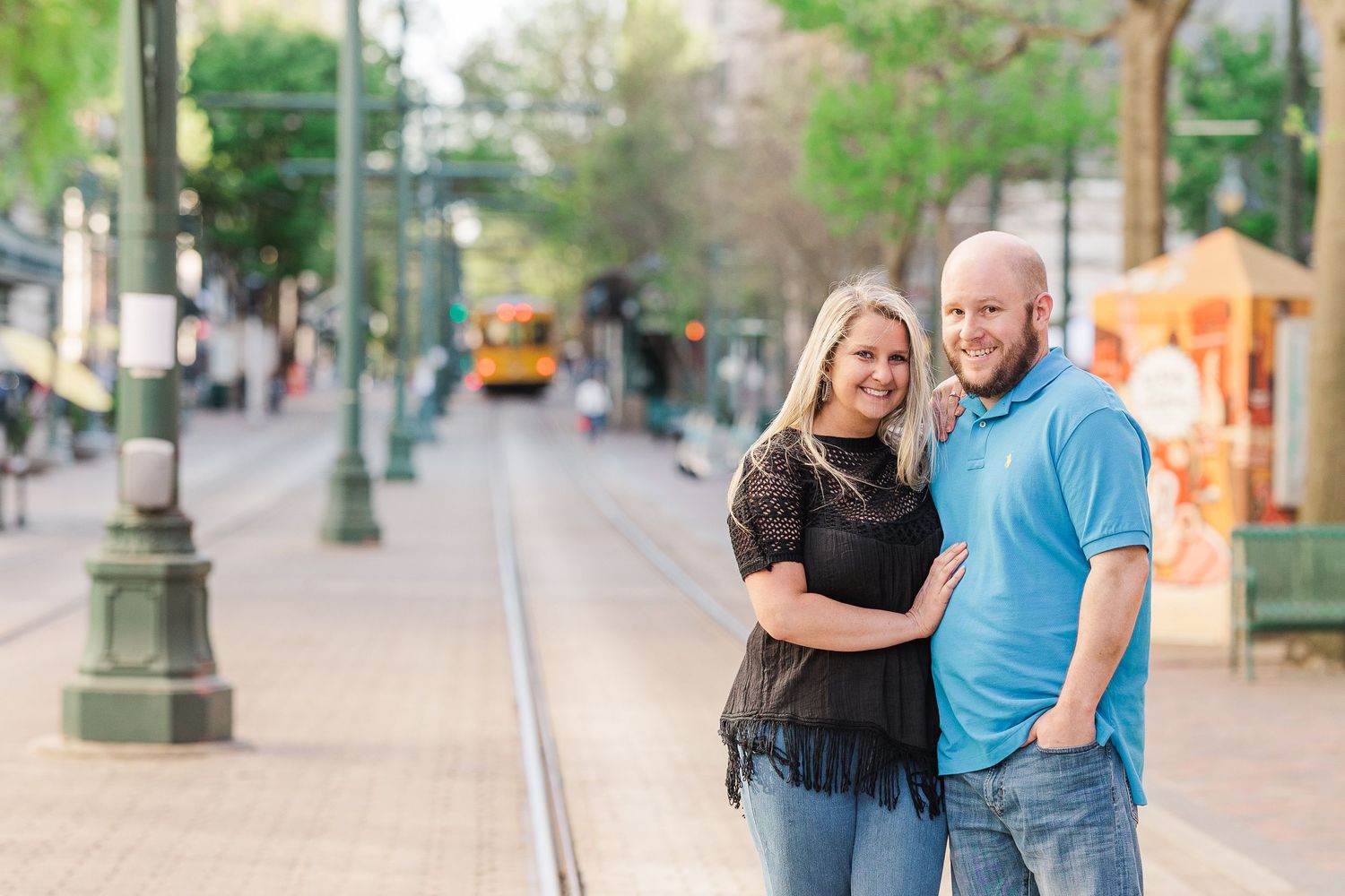 A couple embraces on a tree-lined street with vintage lampposts and trolley tracks.