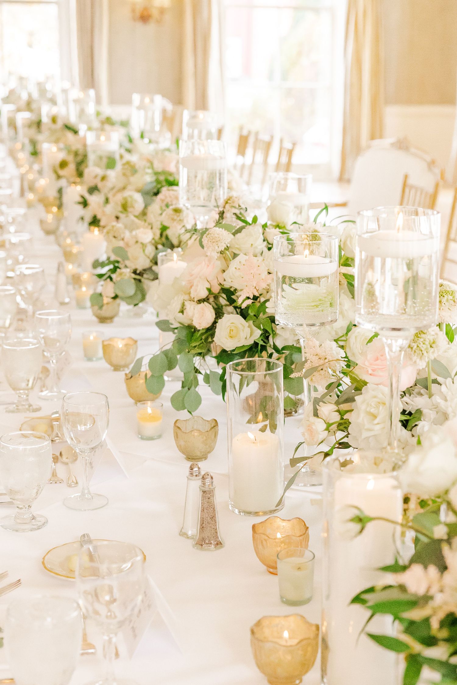 Long wedding table with white floral centerpieces, candles, and gold accents running down white tablecloth.