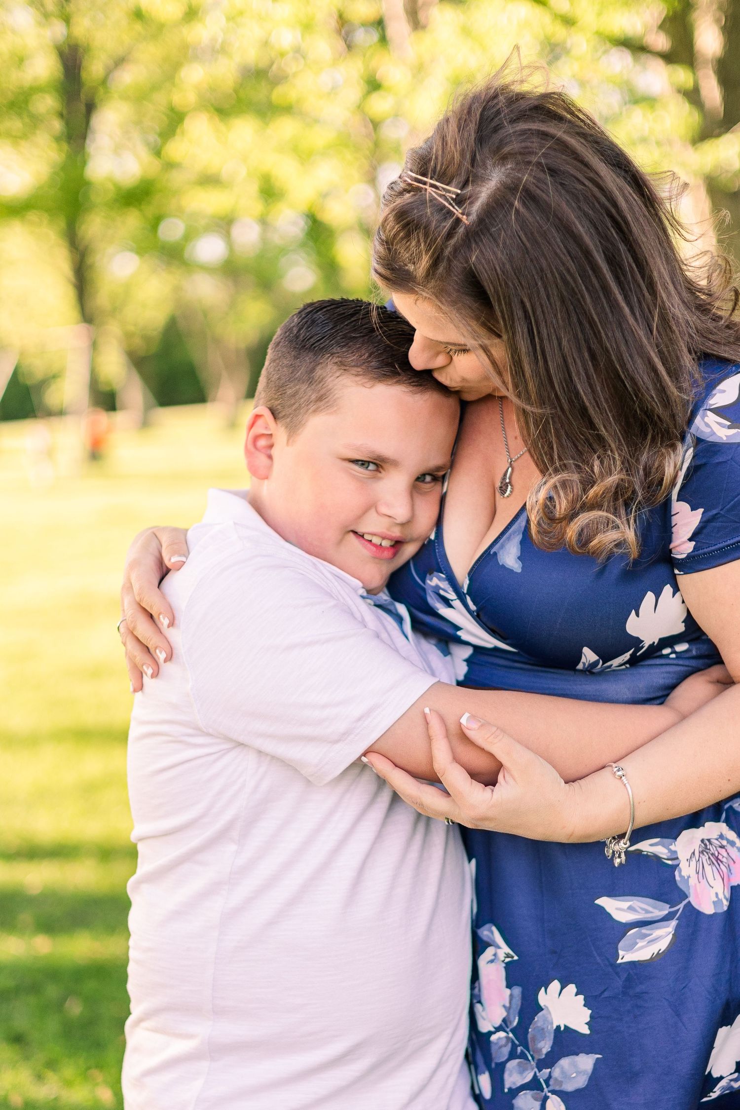 Loving embrace captured in natural sunlight with a blue floral dress visible against green background.