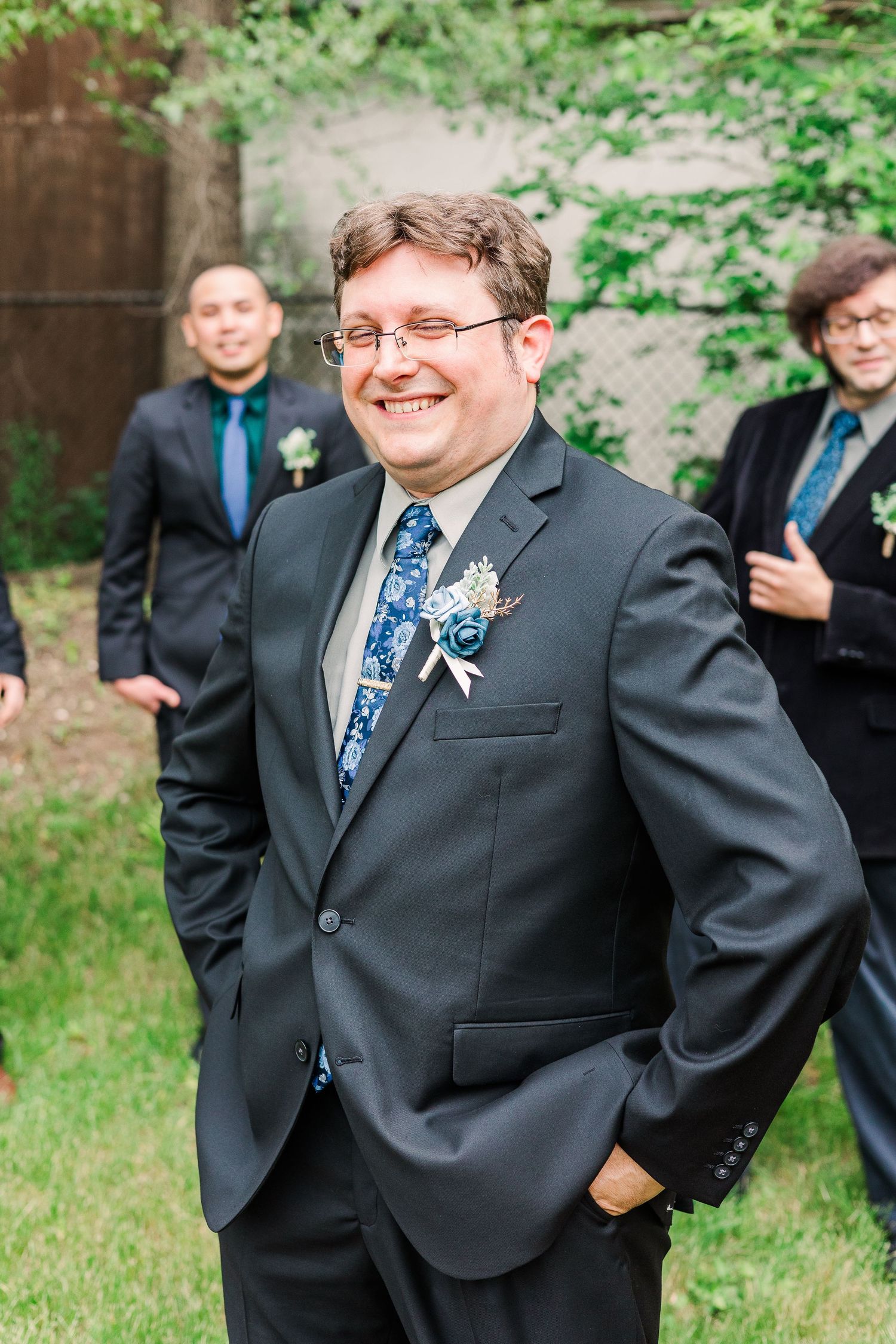 Wedding party member in formal attire smiling outdoors with groomsmen in background.