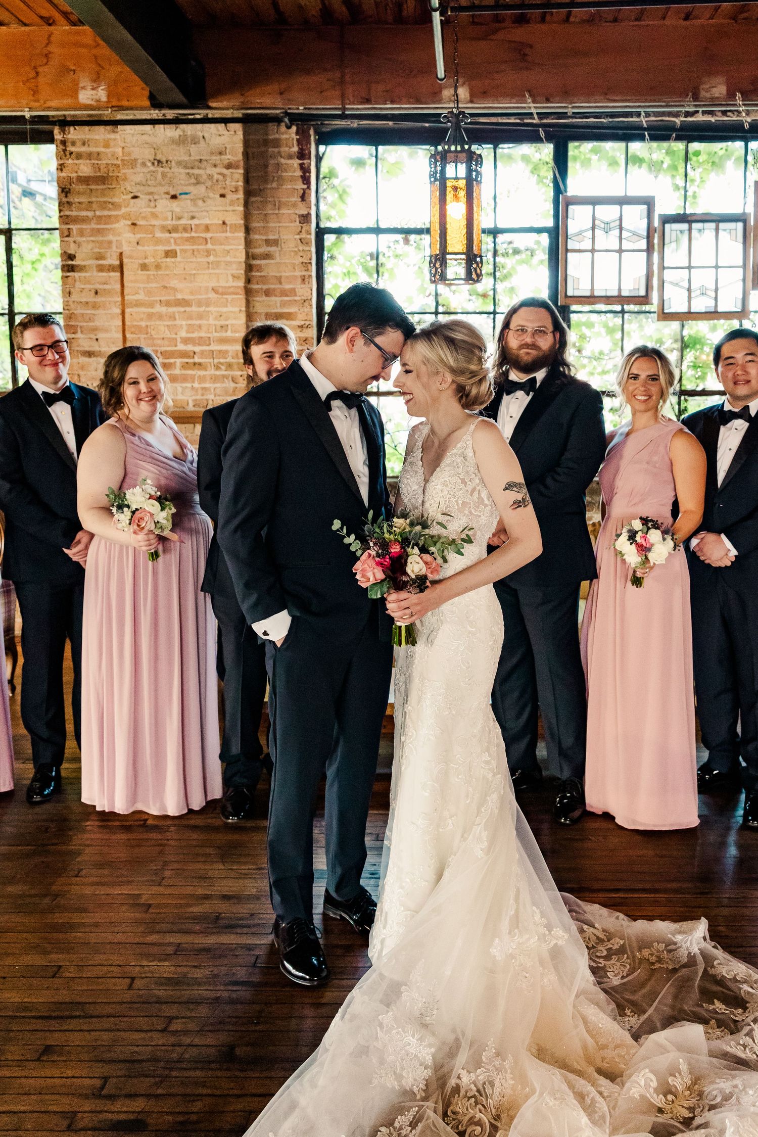 Romantic wedding moment in rustic brick venue with bridal party in pink dresses and black tuxedos.