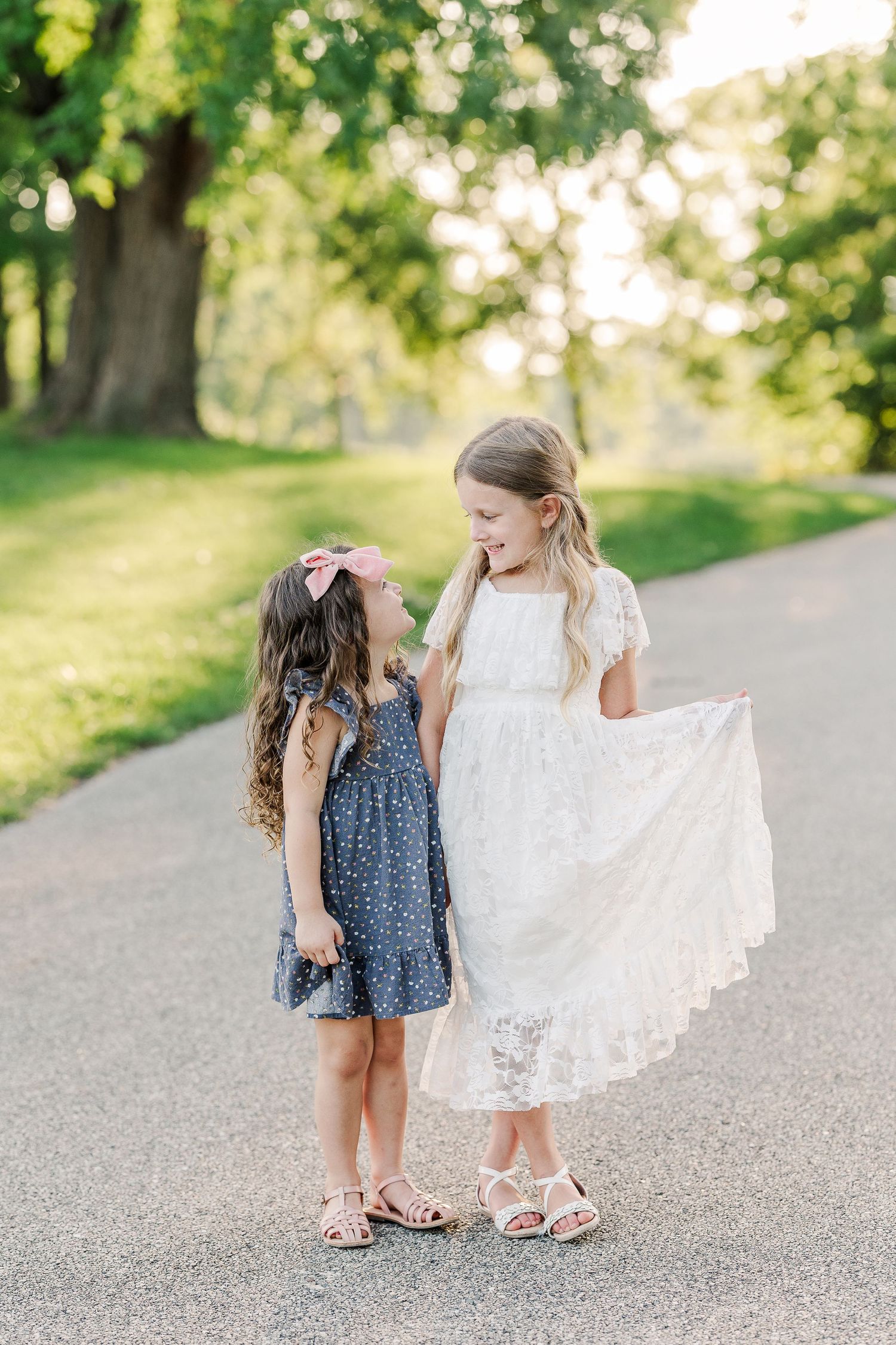 Two children in dresses share a sweet moment on a tree-lined path during golden hour.