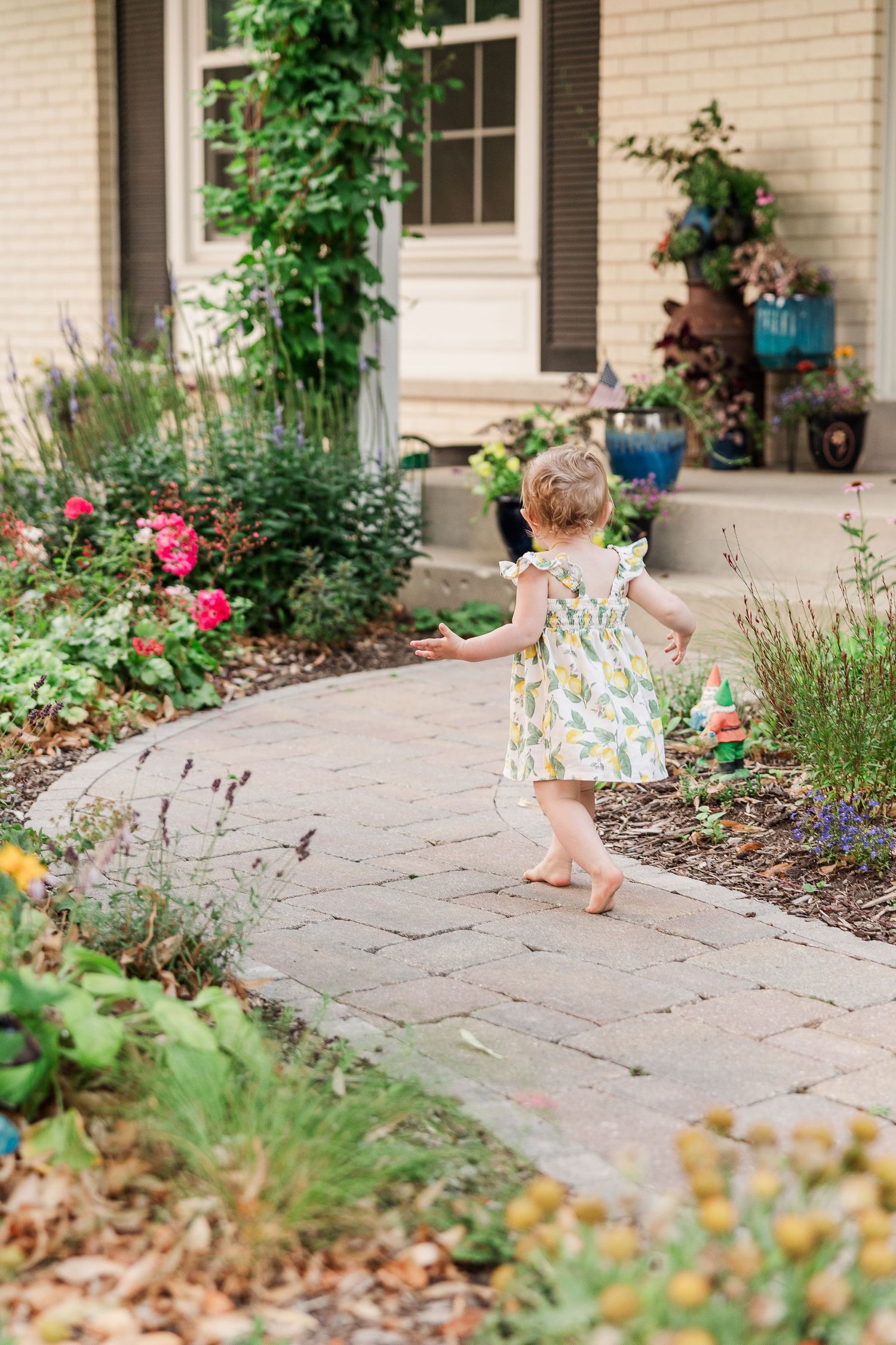 A toddler wearing a floral dress walks barefoot along a curved garden path surrounded by colorful flowers and greenery.