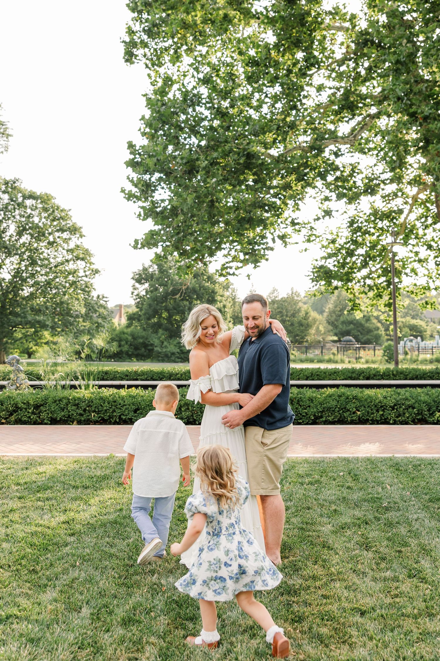 Family of four in a lush green park, parents embracing as two young children run toward them.