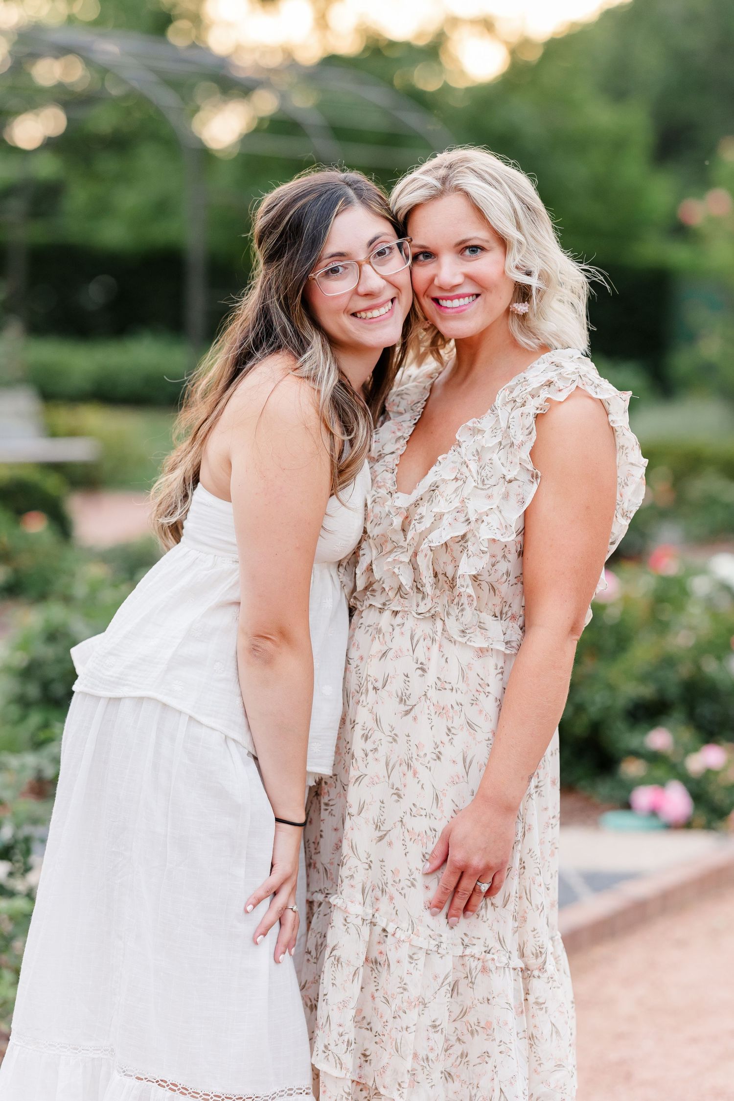 Two smiling women in floral and white dresses pose together in a beautiful garden setting at golden hour.