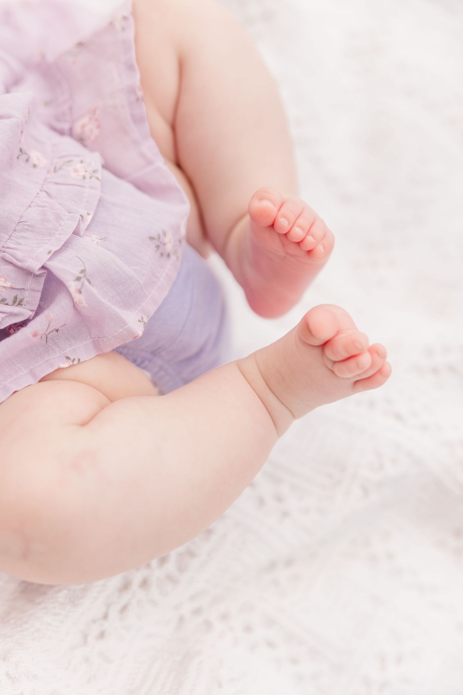 Close-up of tiny newborn baby feet and legs in a soft purple outfit on a white blanket.