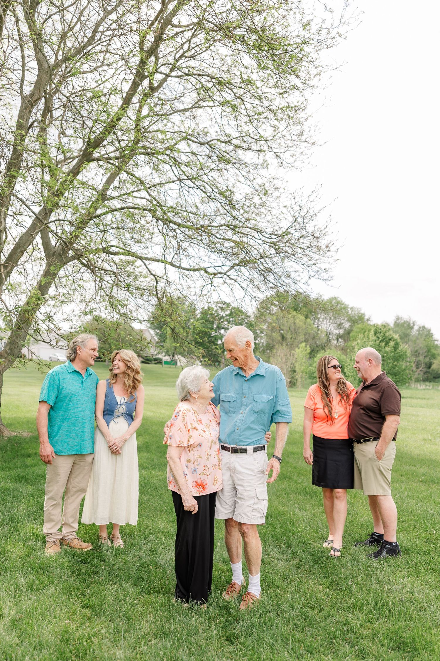 Six family members gather outdoors on green grass beneath a tree, smiling and conversing during a spring family portrait session.