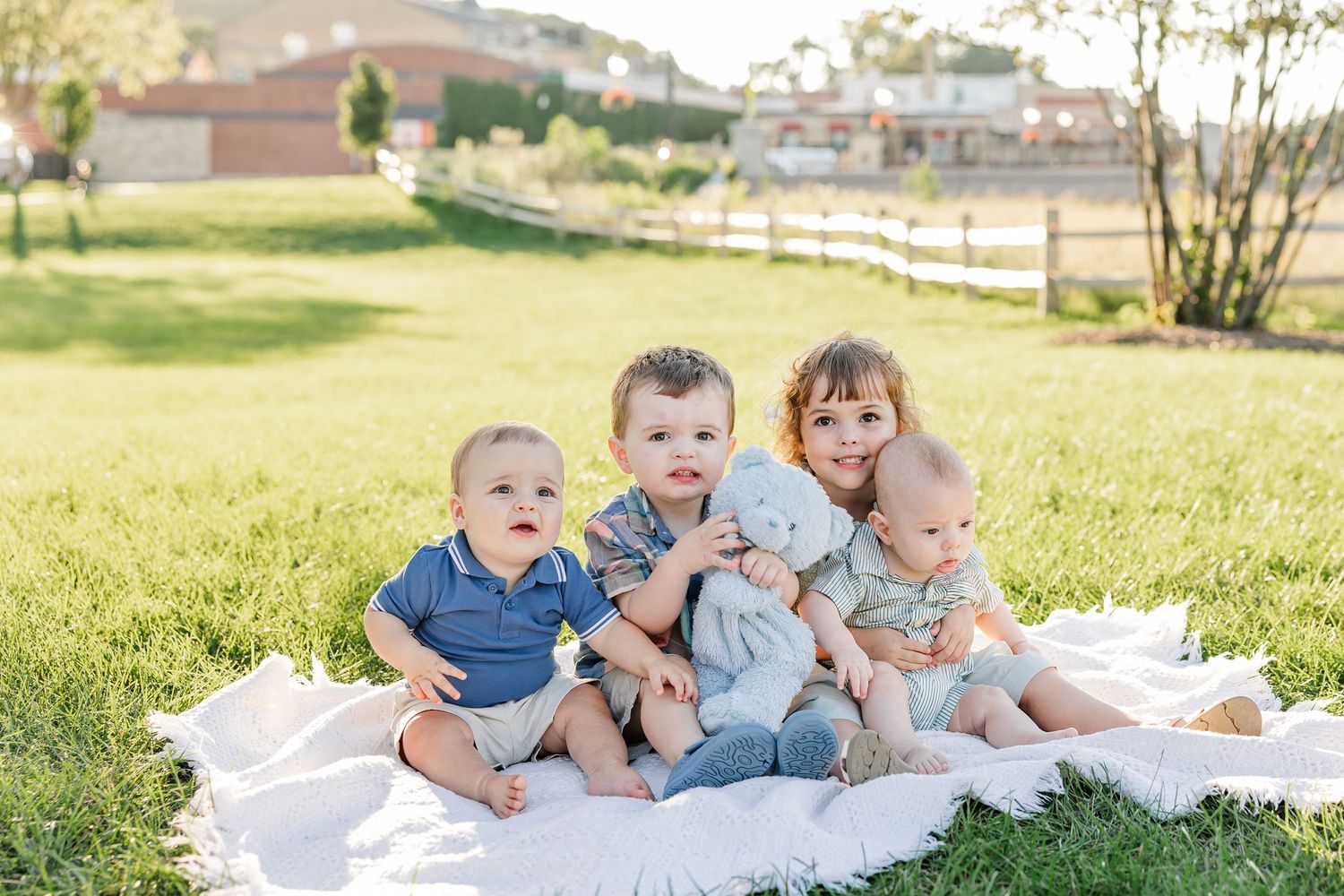 Four young children sit together on a white blanket on green grass, posing for an outdoor family photo.