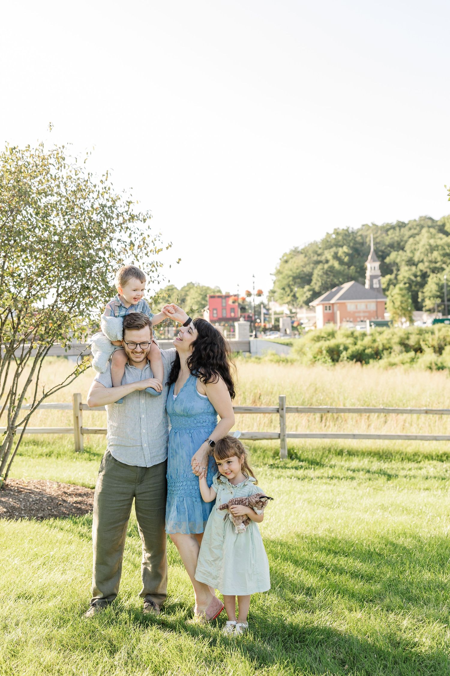 A happy family of four poses outdoors on green grass, with a boy on dad's shoulders and a girl in a light blue dress.