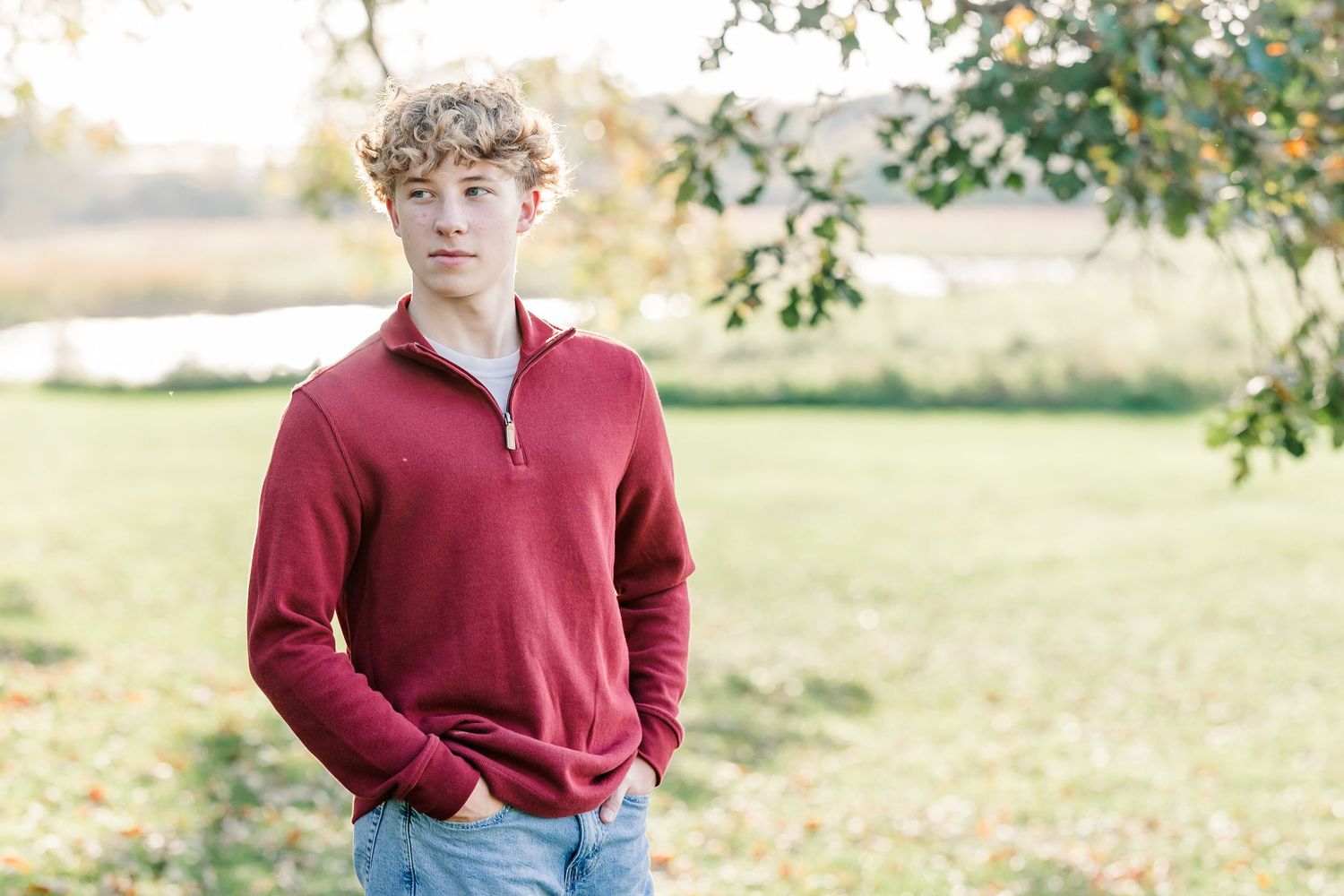 Young man with curly blond hair wearing a red quarter-zip pullover and jeans stands outdoors with hands in pockets in a sunlit grassy park.