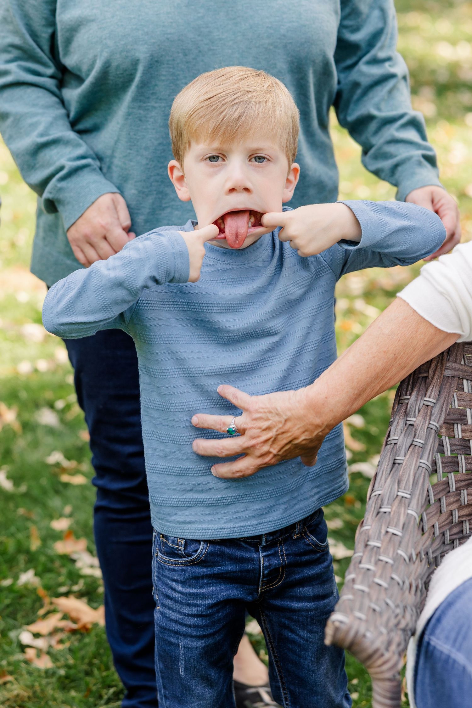Playful blonde boy sticking out tongue in a blue shirt outdoors in a park.