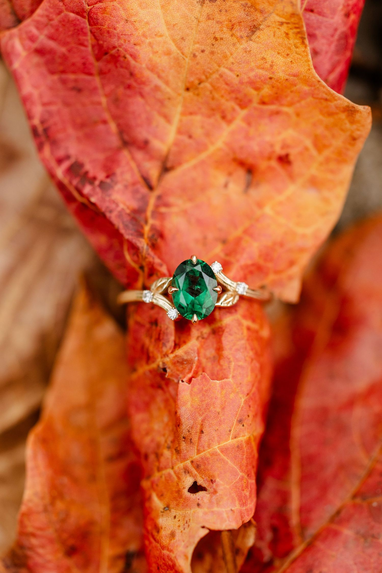 Oval green emerald engagement ring with diamond accents resting on vibrant red and orange autumn leaves.