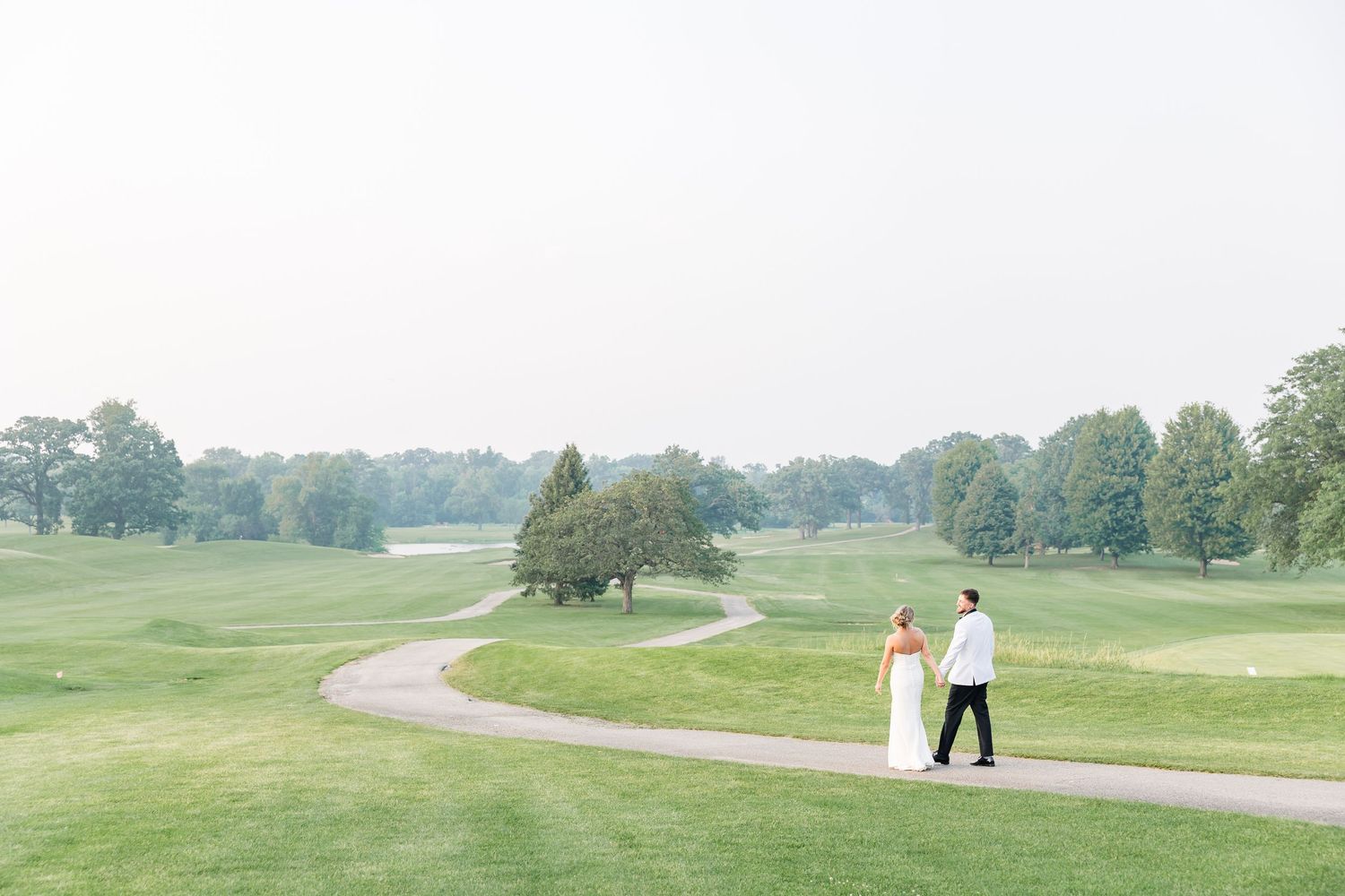 Bride and groom walk hand in hand along a winding path through a lush green golf course.
