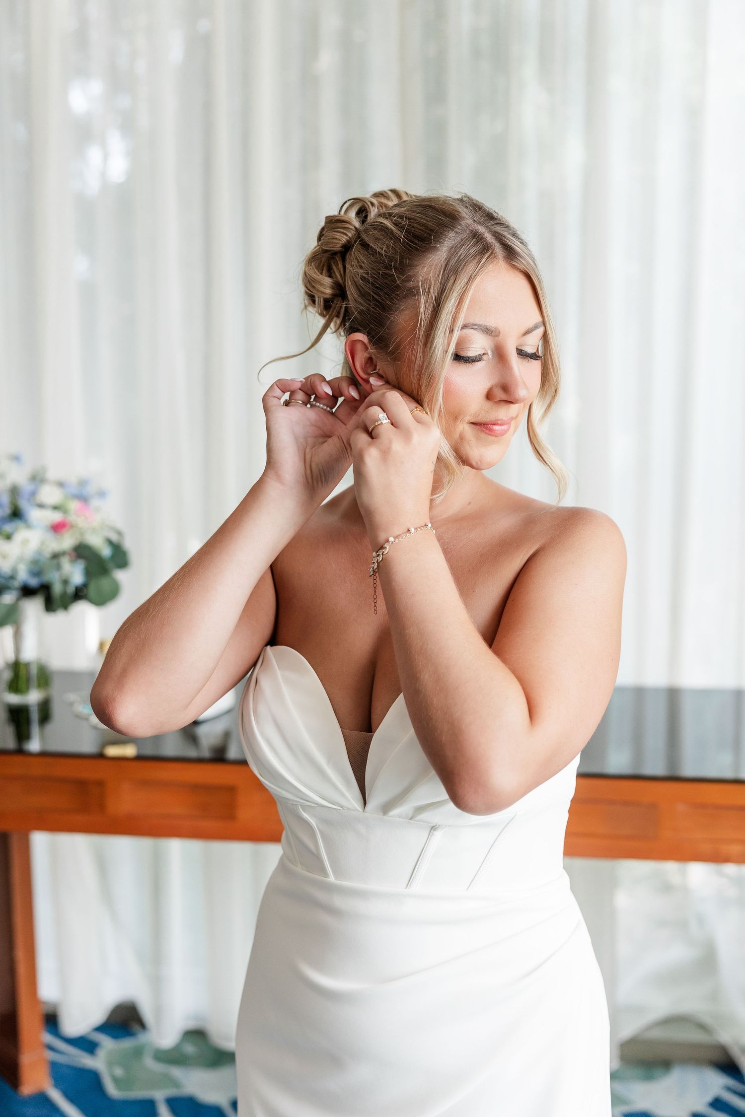 Blonde bride in white sweetheart gown putting on earrings while getting ready near a window.