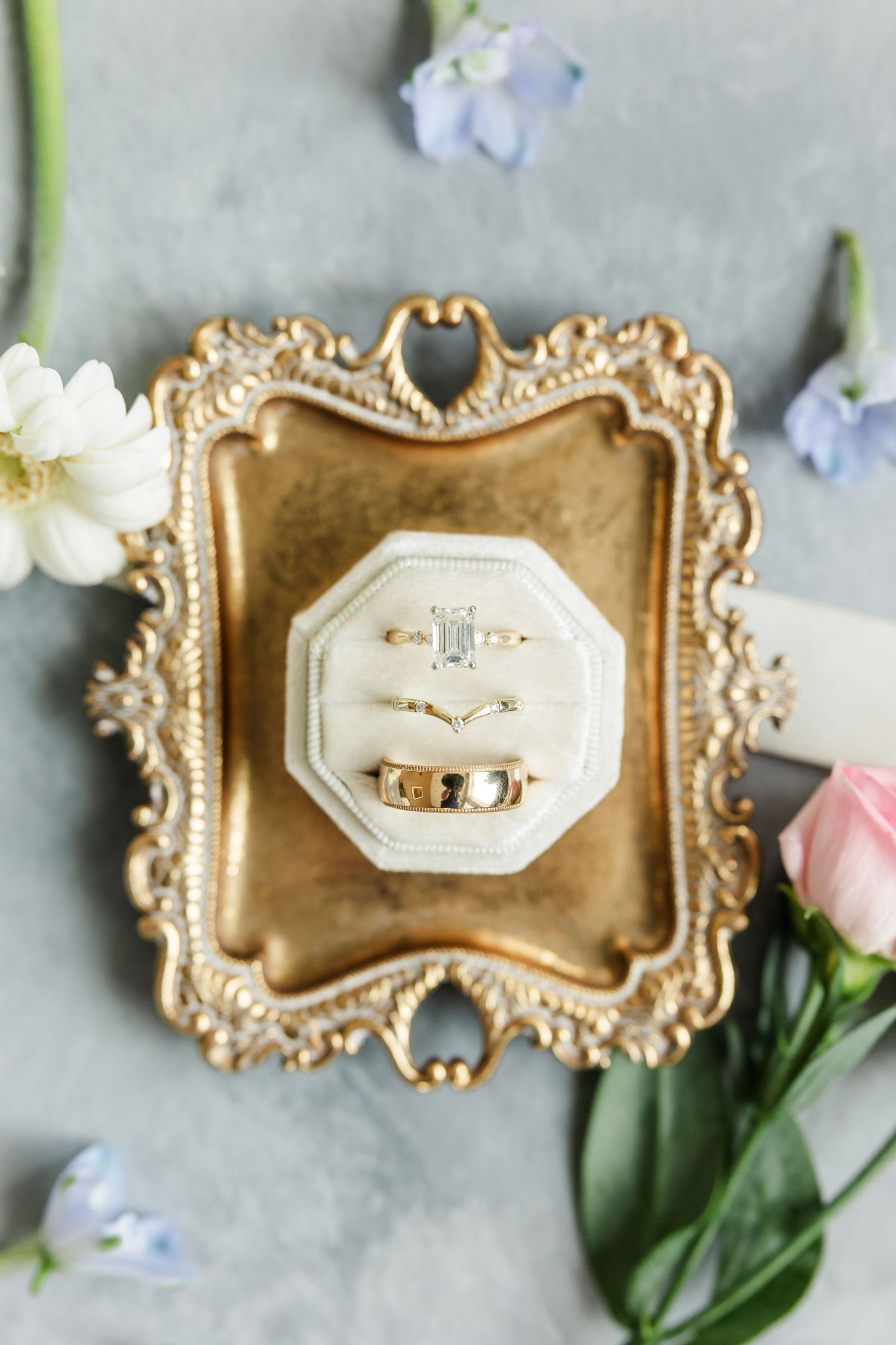 Gold vintage tray with velvet ring box holding diamond engagement ring and wedding bands surrounded by delicate flowers.