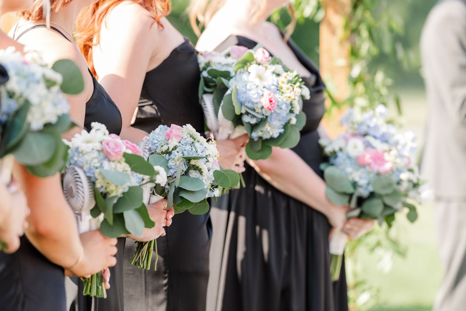 Bridesmaids in black dresses hold bouquets of blue hydrangeas, pink roses, and eucalyptus greenery.