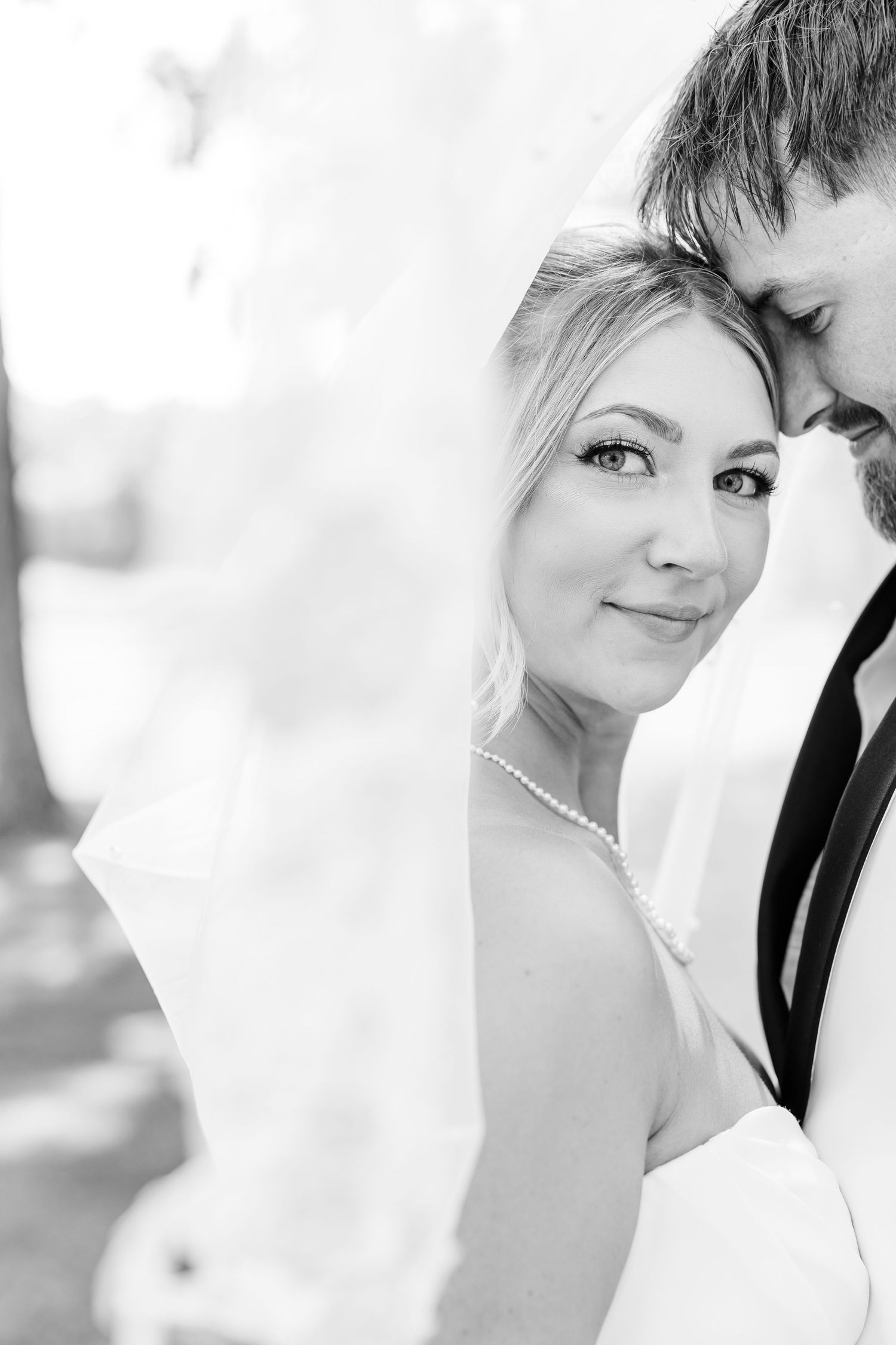 Black and white portrait of a smiling bride in veil and pearl necklace embracing her groom at wedding.