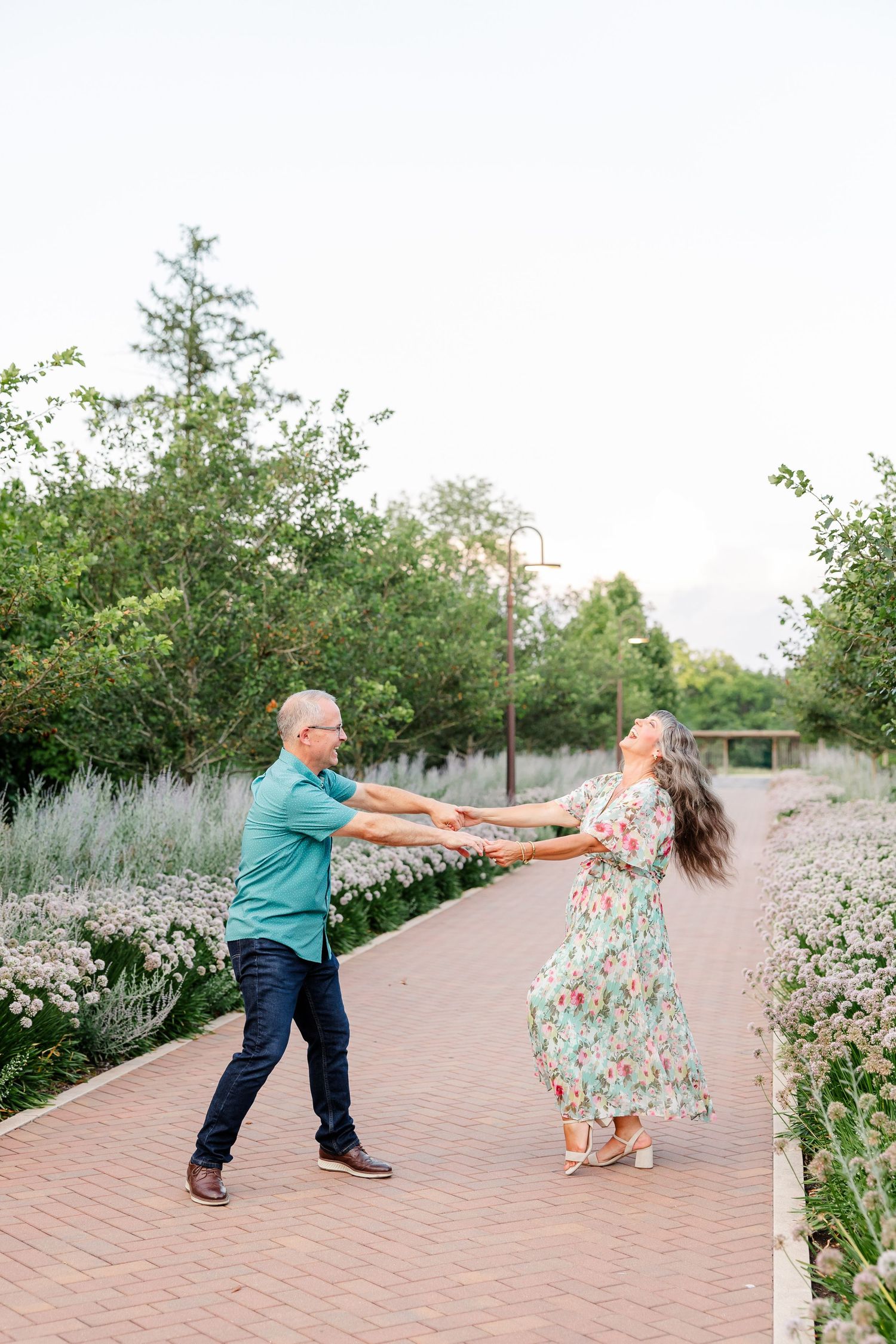 Couple dancing joyfully on a brick garden path, man in teal shirt, woman in floral dress, hands outstretched.