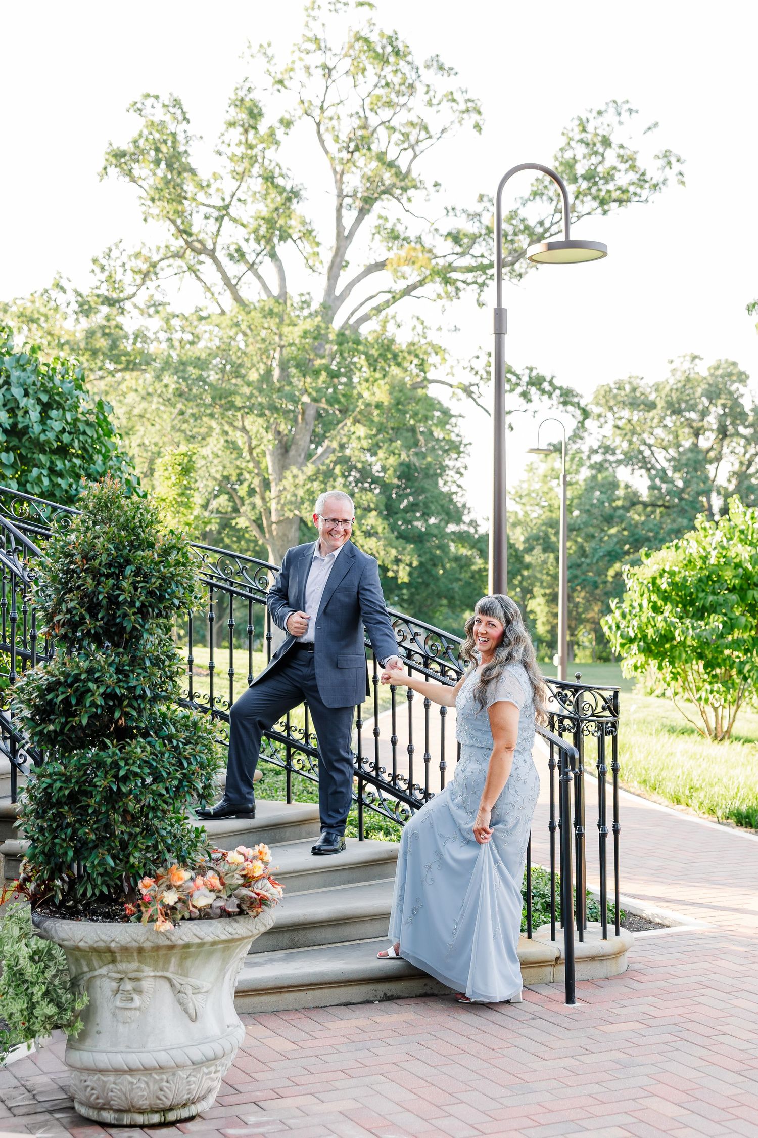 Couple in formal attire posing on outdoor stone steps with iron railing, lush greenery and decorative urn in background.