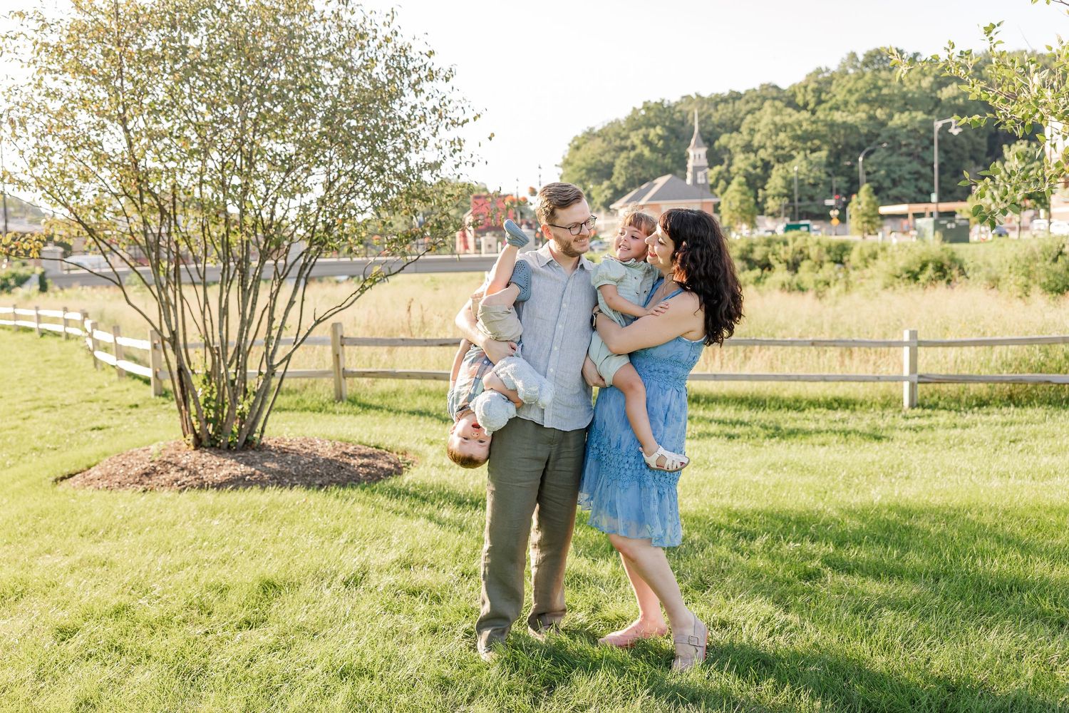 A joyful family of four plays outdoors on green grass, with parents holding young children near a wooden fence.