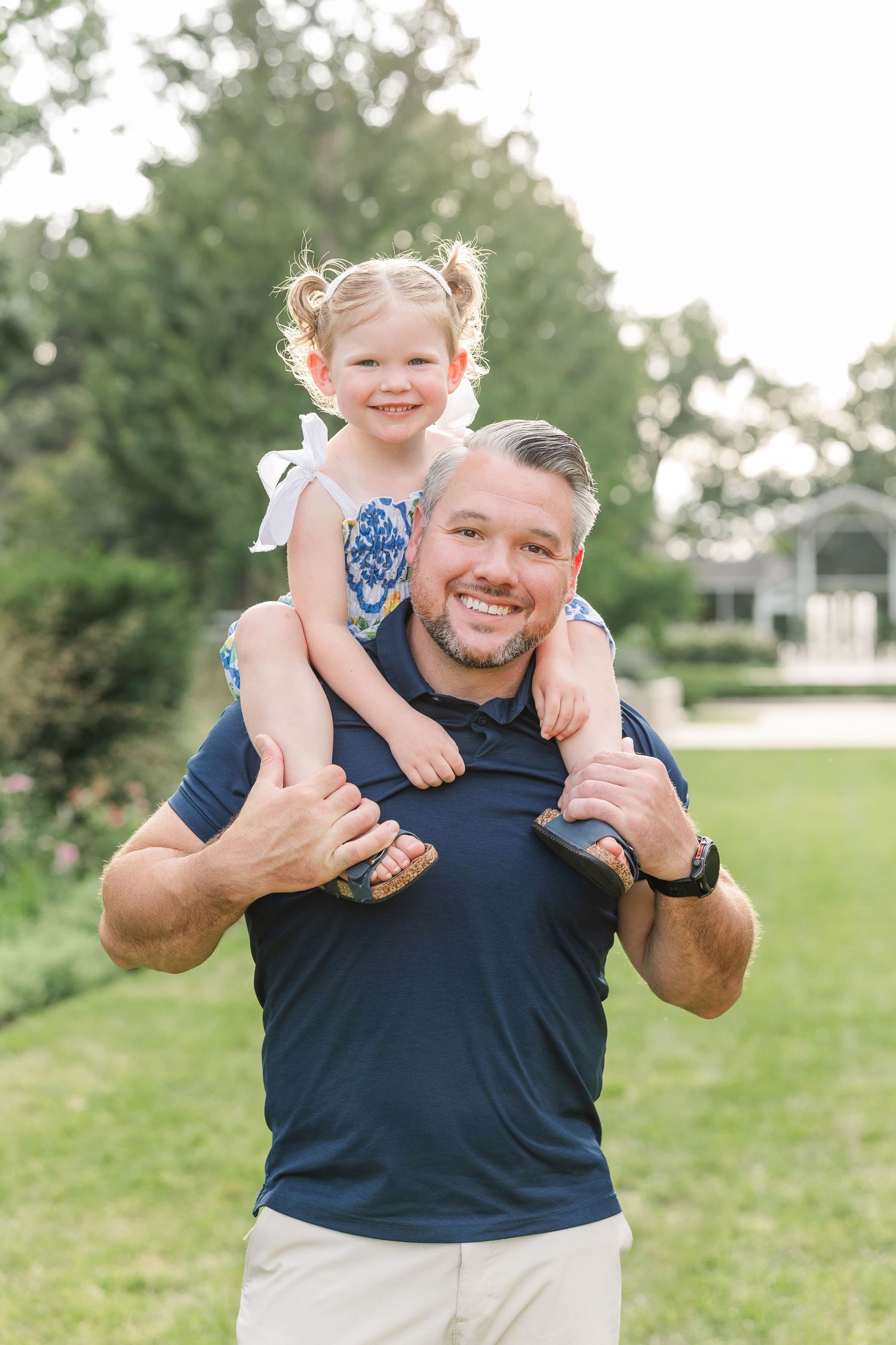 Smiling dad with young blonde girl sitting on his shoulders outdoors.