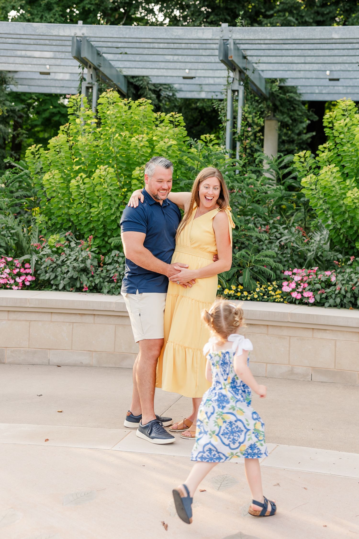 Laughing pregnant couple in a garden watches their young daughter running toward them in a floral dress.